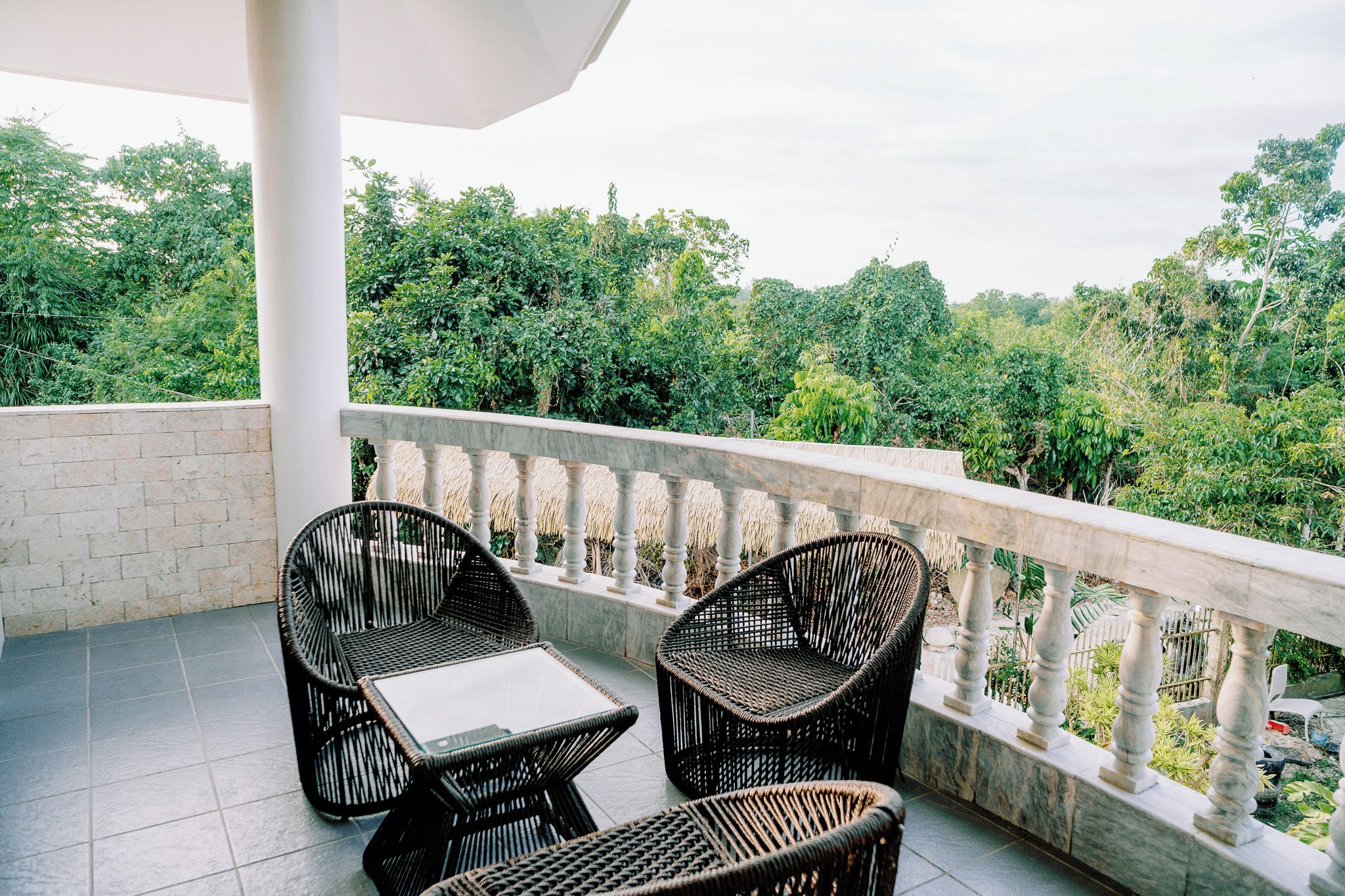 A balcony with two wicker chairs and a small table, surrounded by a white balustrade, overlooking a lush green landscape.