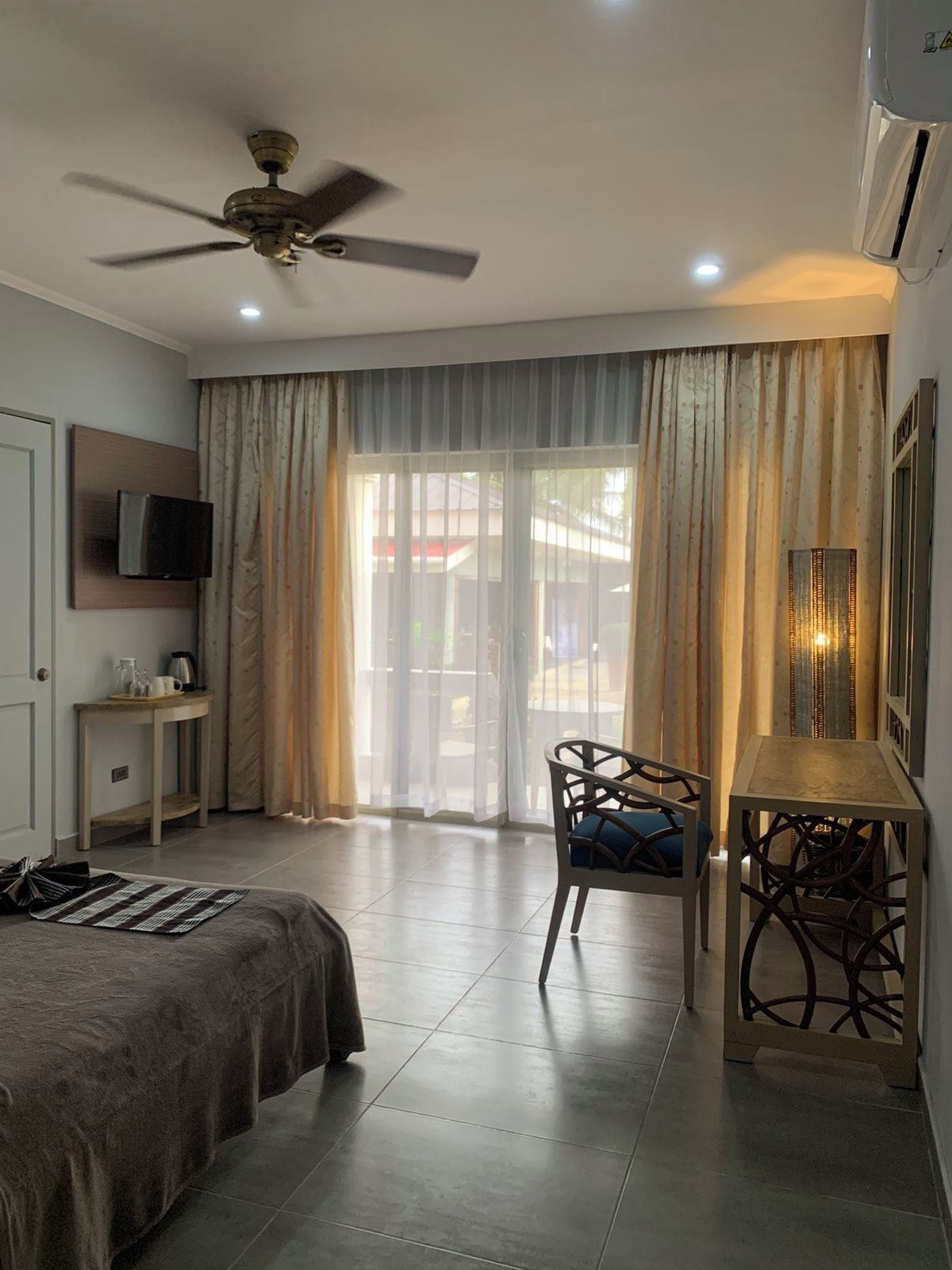 Une chambre d’hôtel équipée d’un lit, d’un bureau, d’une chaise, d’un ventilateur de plafond et d’un téléviseur fixé au mur.