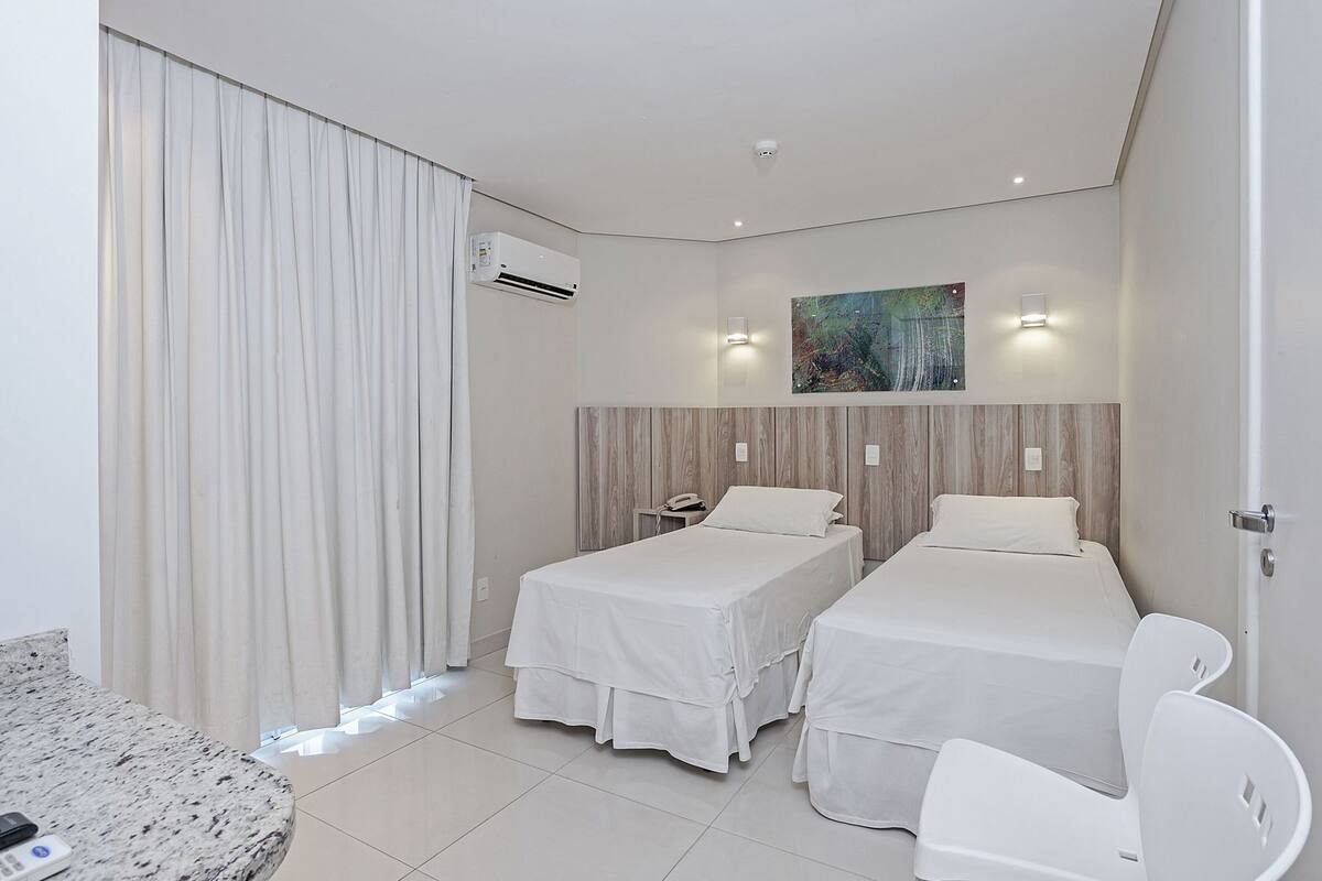A hotel room with two beds, a painting on the wall, an air conditioning unit, and a small table with a chair.