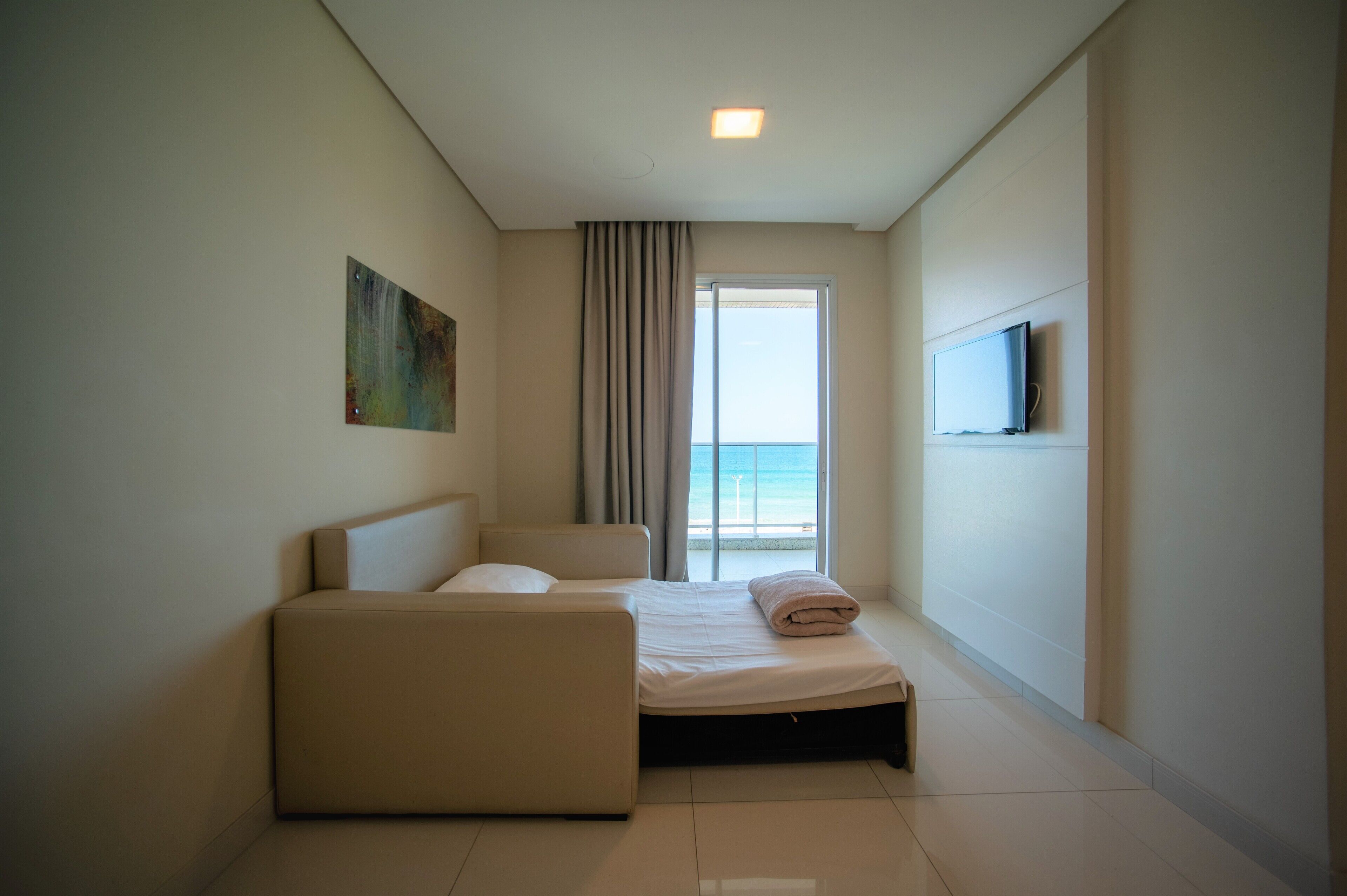 A hotel room with a bed, a sofa, a TV mounted on the wall, and a large window with a view of the sea.