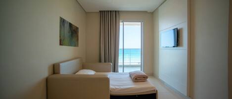 A hotel room with a bed, a sofa, a TV mounted on the wall, and a large window with a view of the sea.