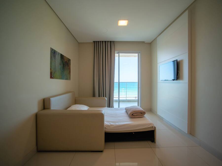 A hotel room with a bed, a sofa, a TV mounted on the wall, and a large window with a view of the sea.