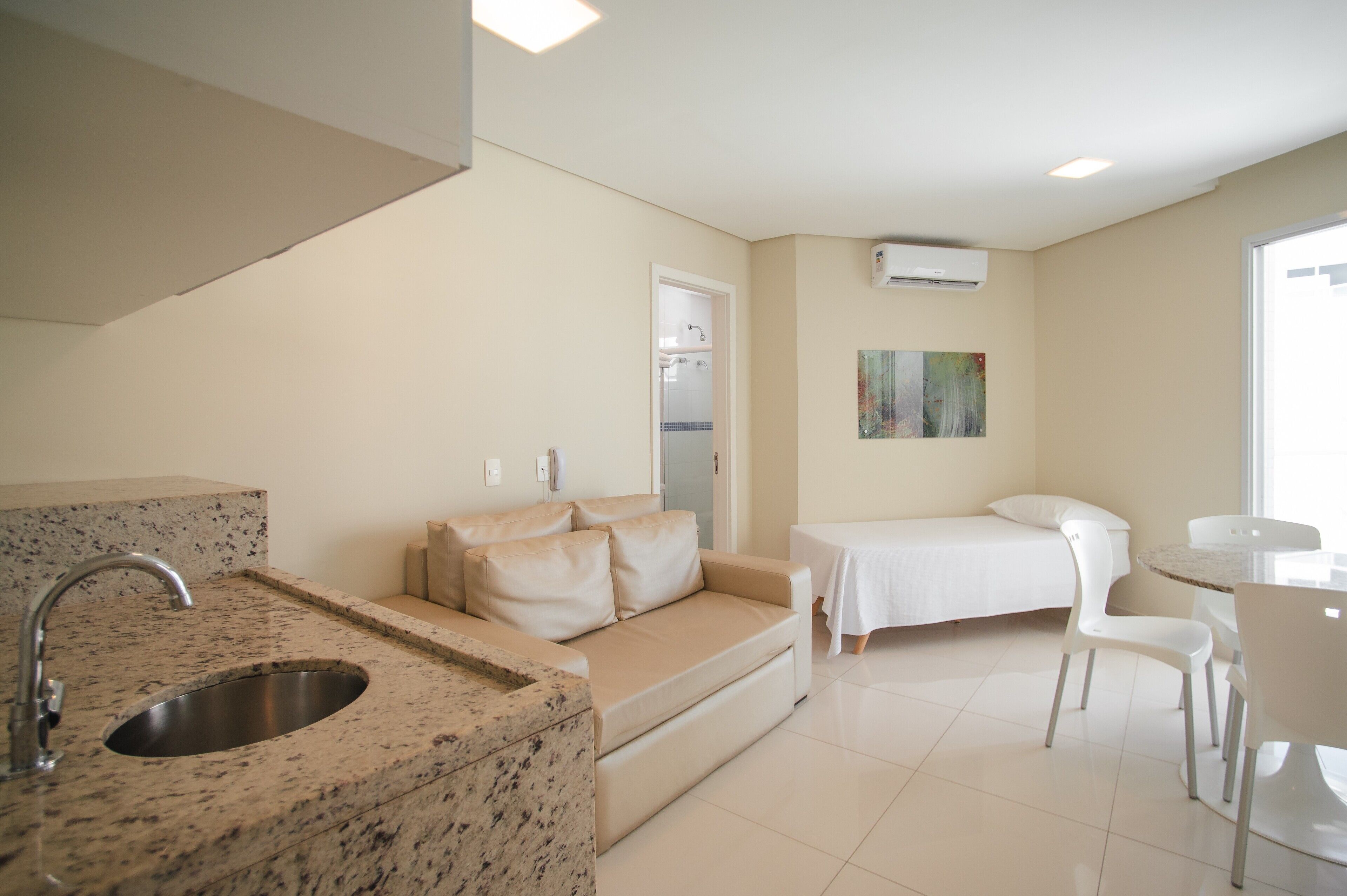 Une cuisine moderne avec un plan de travail et un évier en granit, un canapé beige, une petite table à manger avec des chaises, et un mur blanc sur lequel est installé un climatiseur.