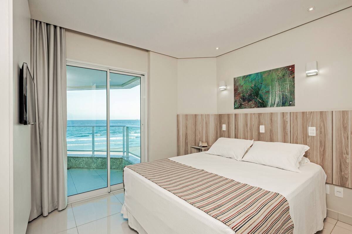 A hotel room with a large bed, a painting on the wall, and a view of the ocean through a sliding glass door.