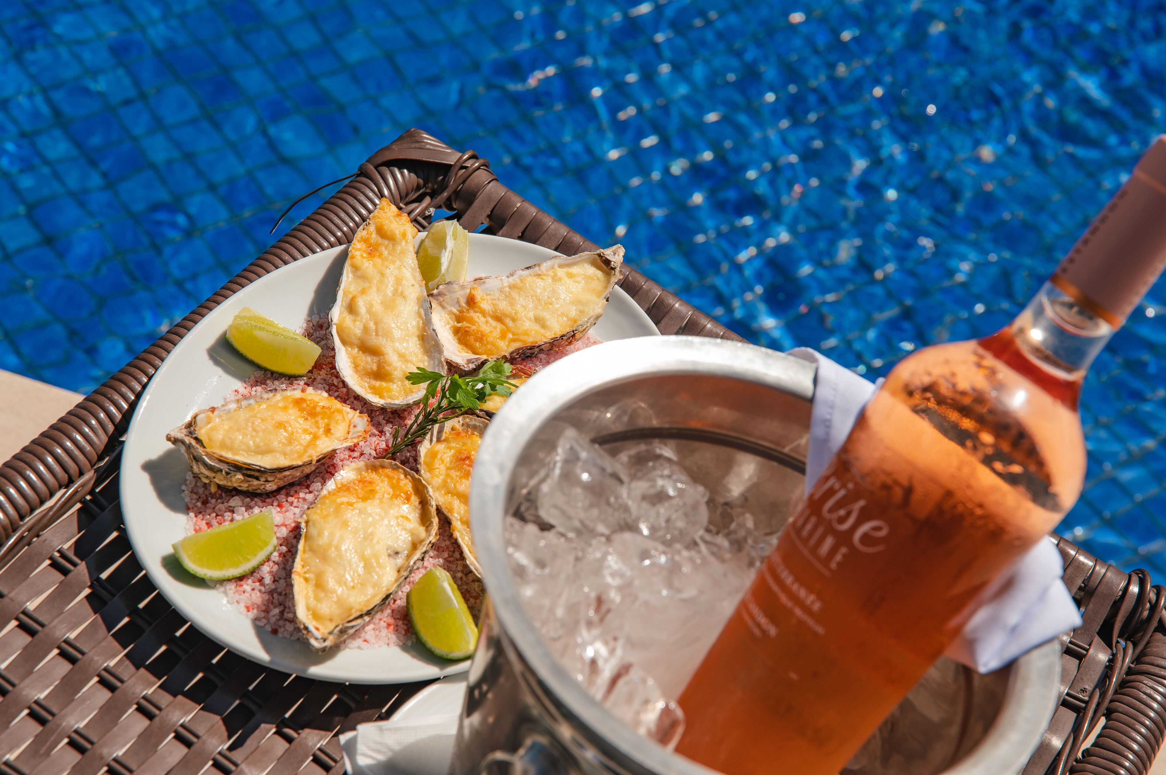 A plate of oysters with lemon wedges on a wicker table by a pool.