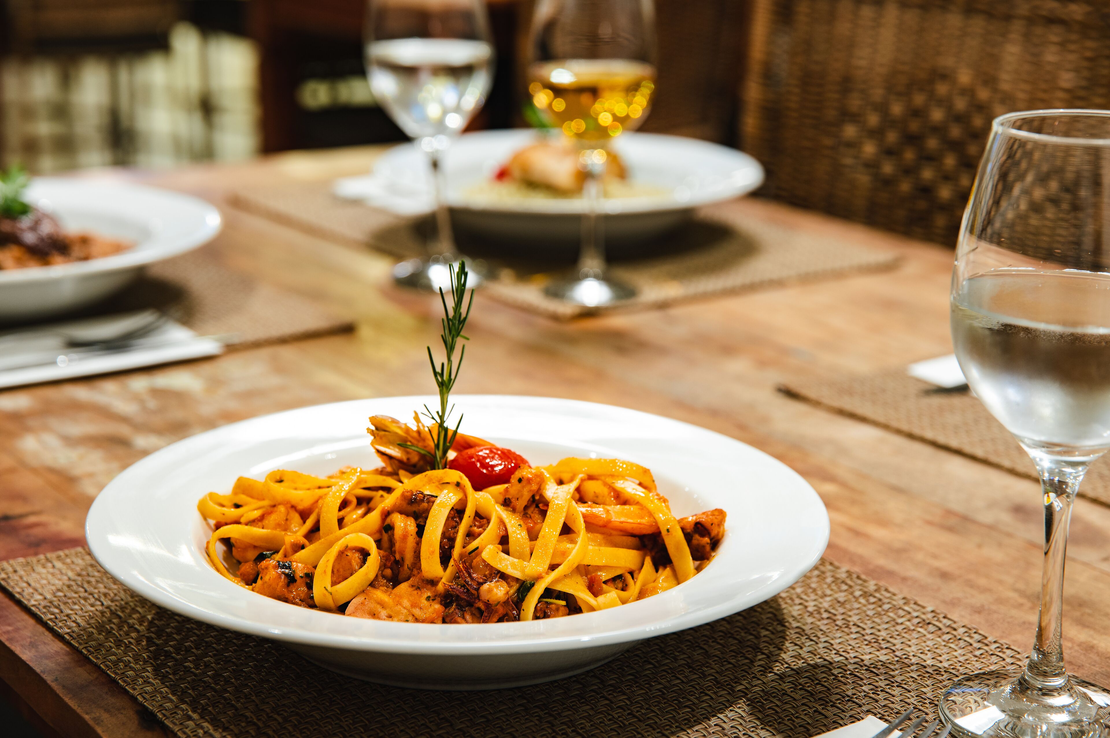 A dining table with a plate of pasta, a glass of water, and a wine glass in the background.
