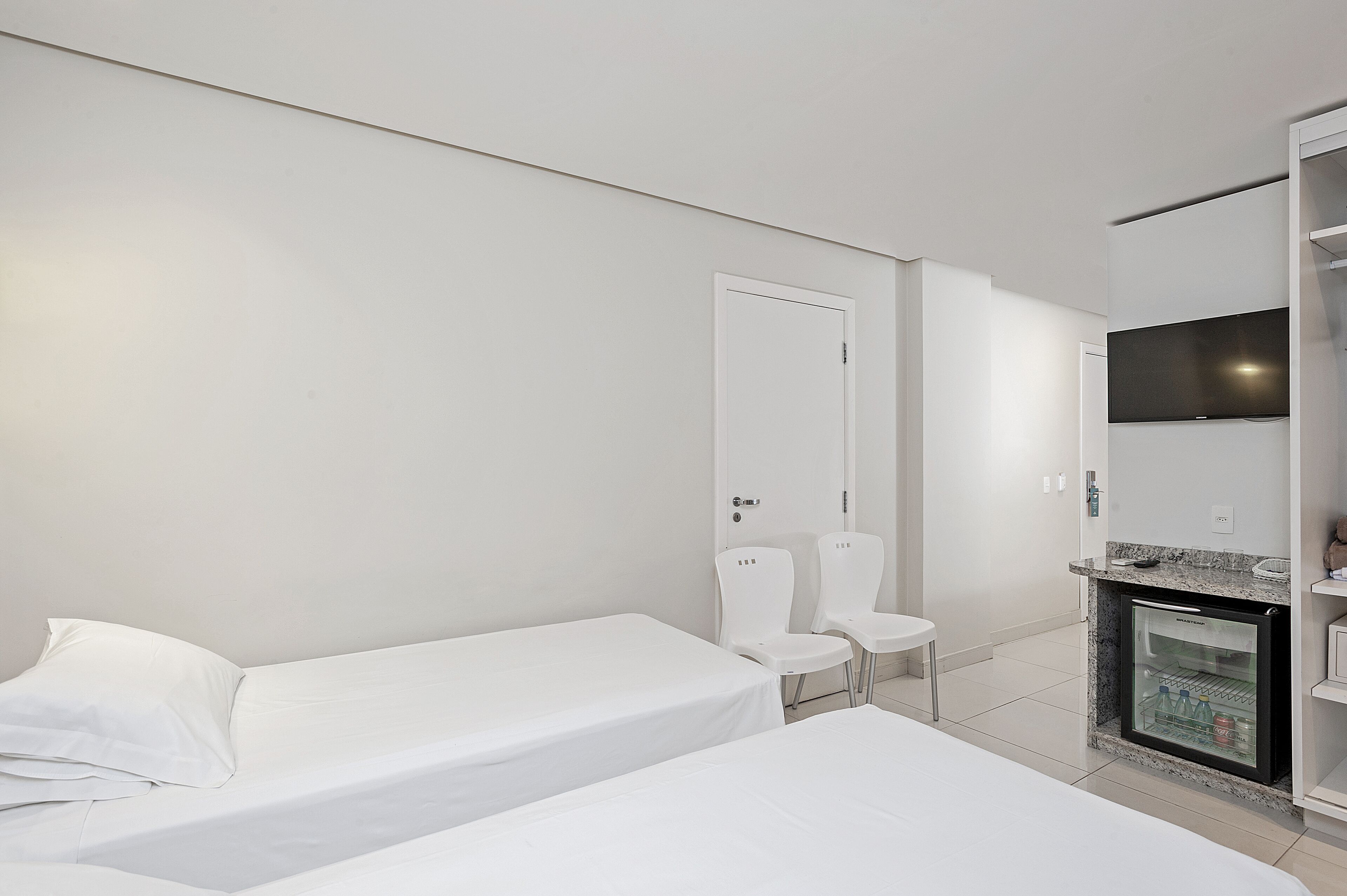 A hotel room with two single beds, a small seating area with chairs, a television mounted on the wall, and a mini-fridge.