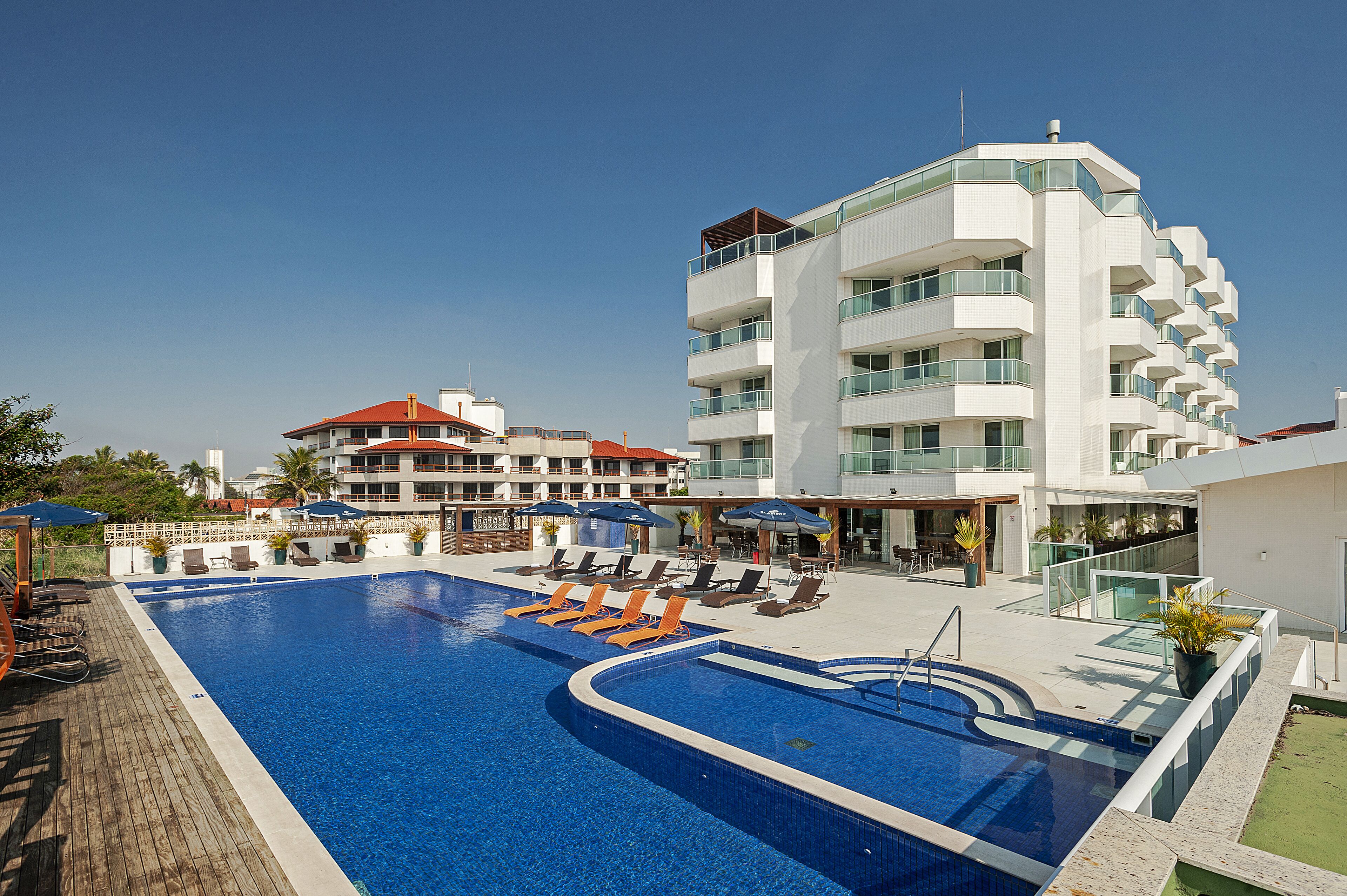 Piscina con sillas de descanso y sombrillas, rodeada por un edificio moderno con balcones.