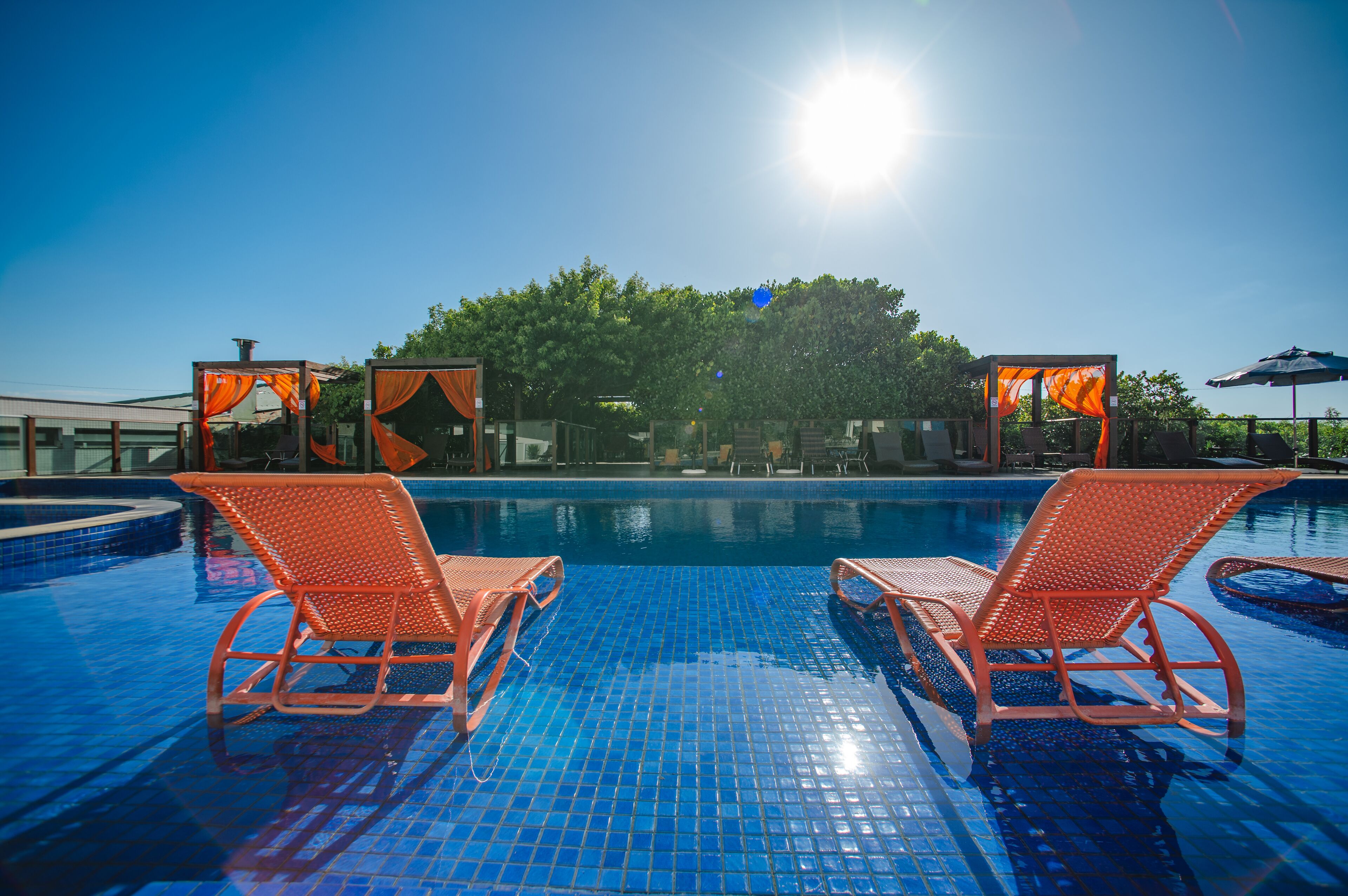 A clear blue pool with two orange lounge chairs facing the water, surrounded by a tiled deck and a covered seating area with orange curtains.