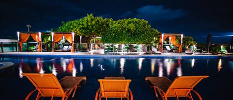 Zona de piscina nocturna con sillones de descanso y un árbol en el centro.