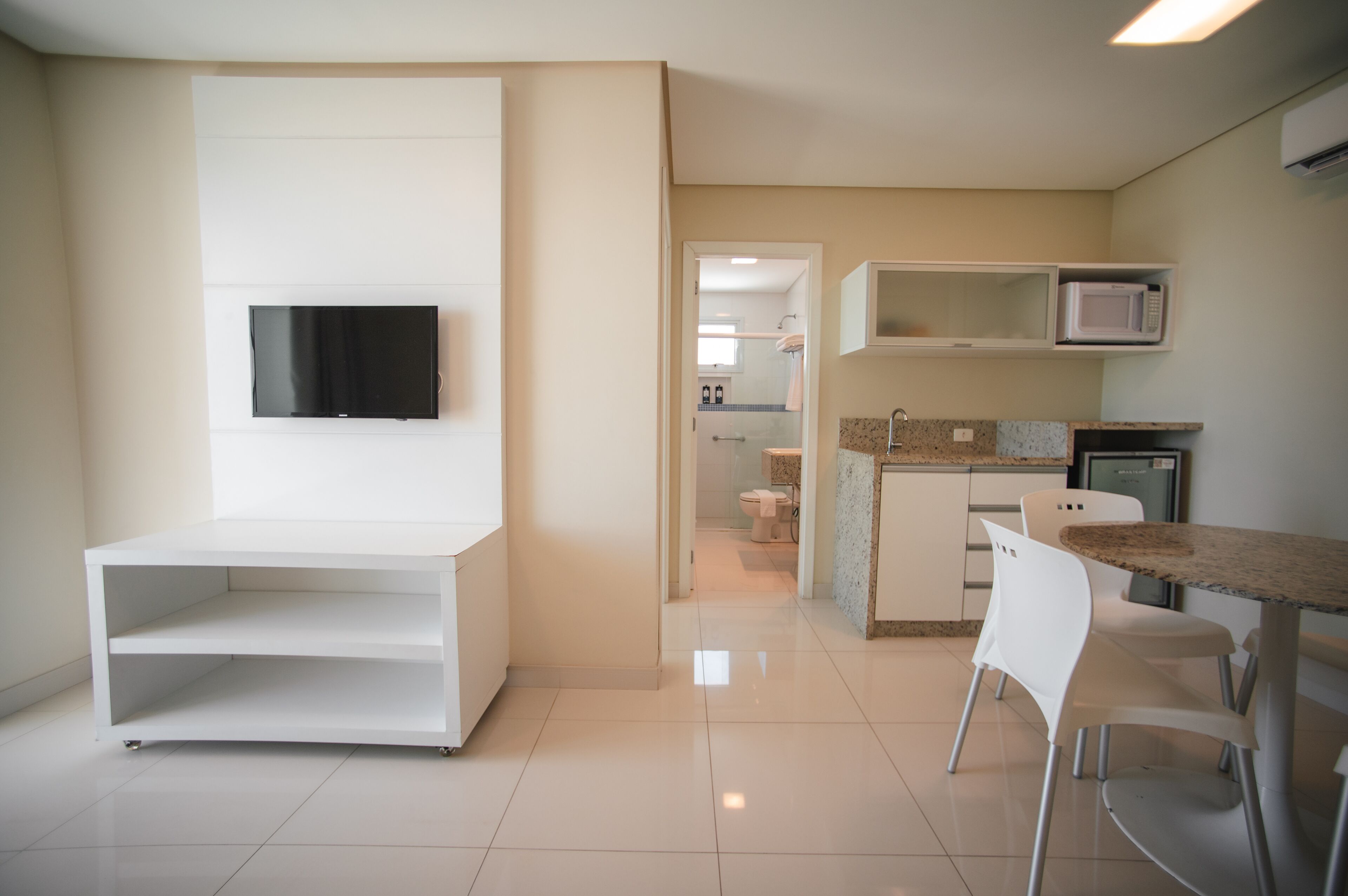 A modern kitchen with a flat-screen TV, white cabinetry, a granite countertop, and a dining area with white chairs.