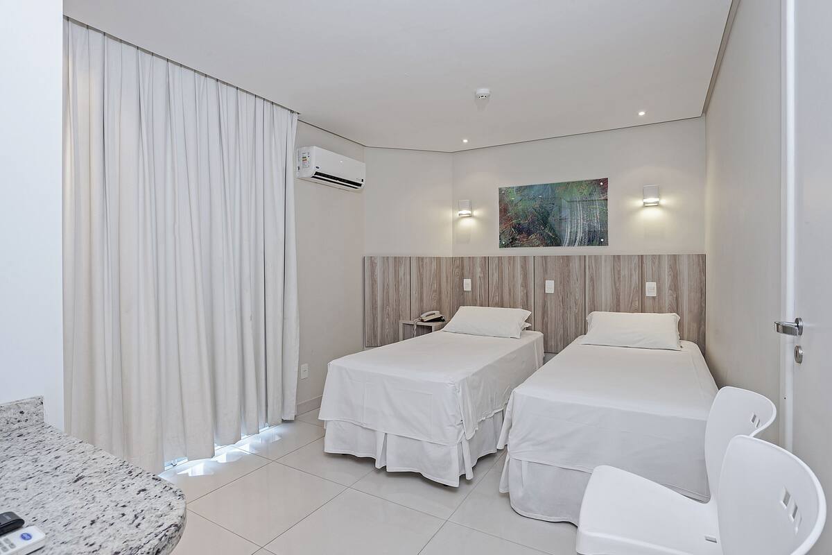 A hotel room with two beds, a desk, and a chair. There is an air conditioning unit on the wall and a painting above the beds.