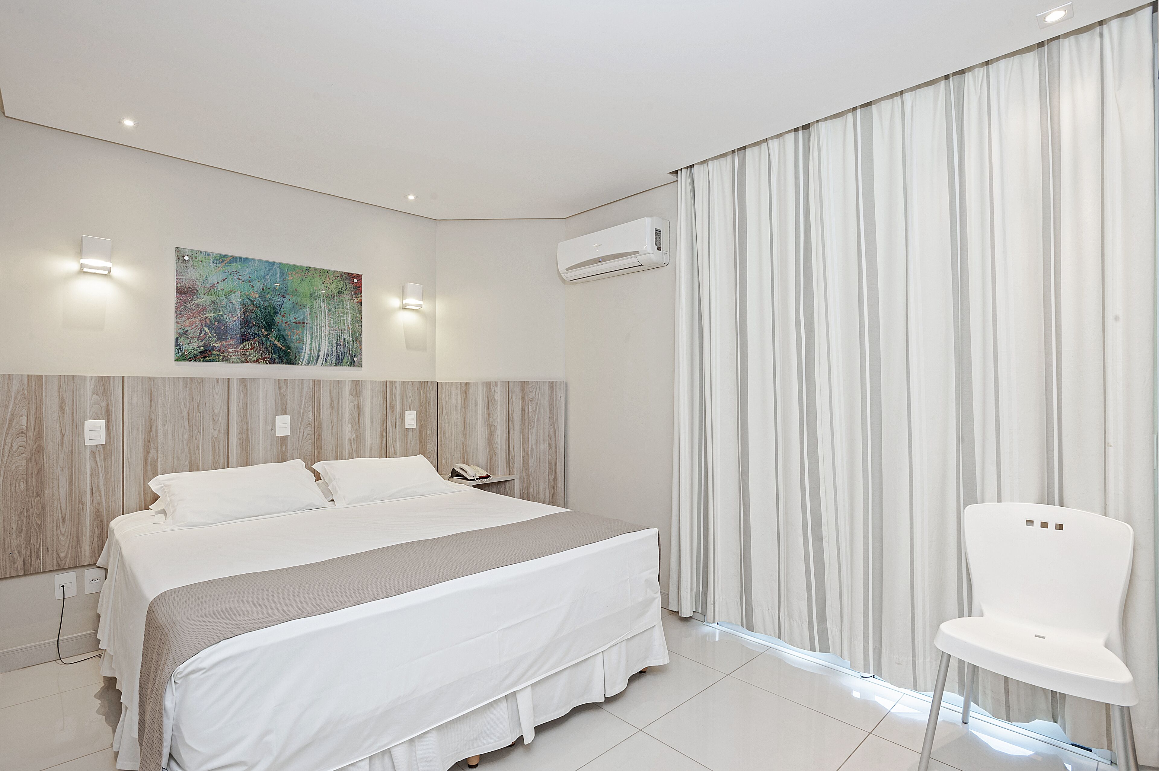 A modern hotel room with a large bed, a white chair, a painting on the wall, and an air conditioning unit.