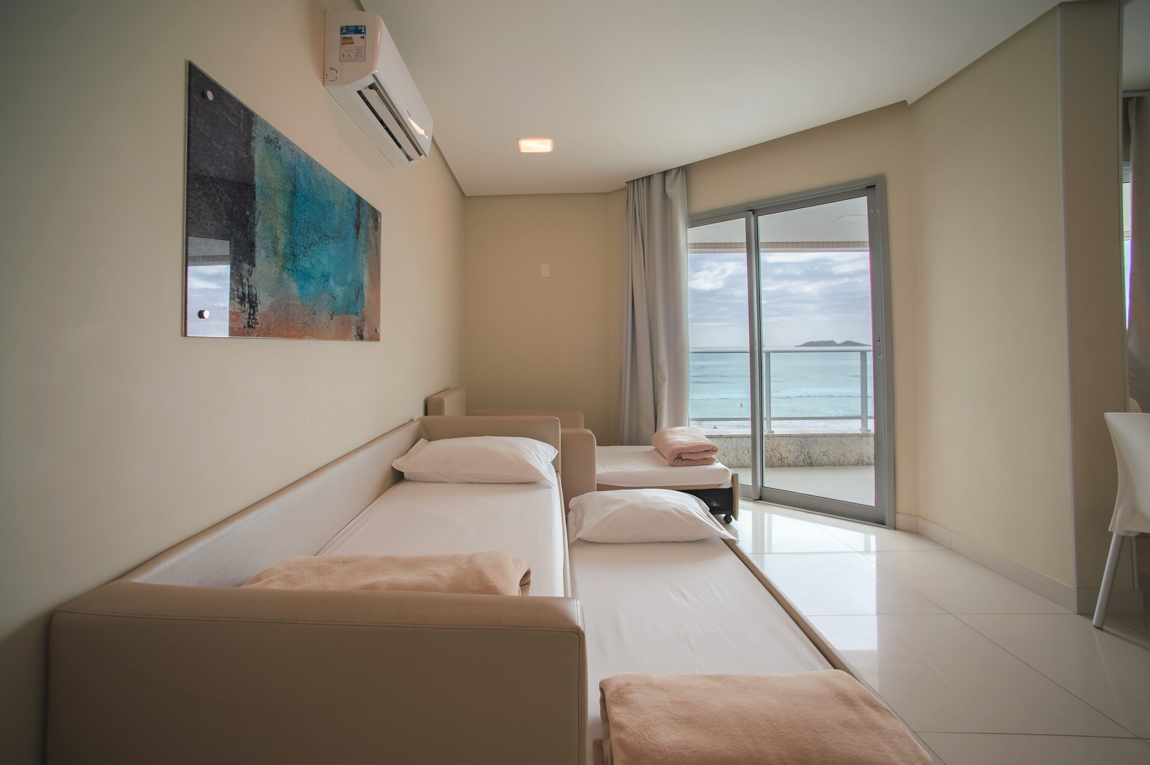A hotel room with three beds, a large window with a view of the sea, and a painting on the wall.
