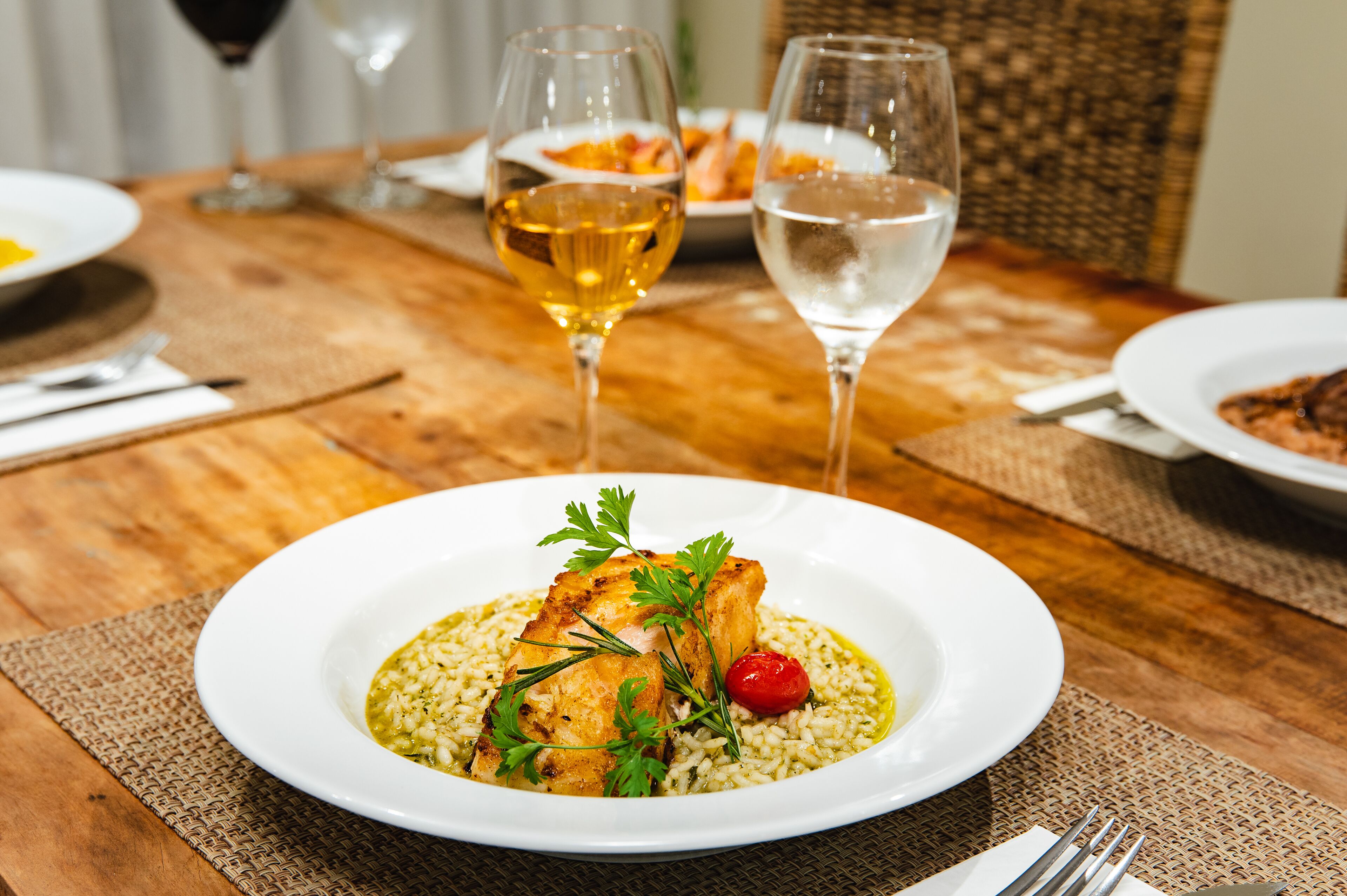 A dining table with a white plate of food, two wine glasses, and a woven placemat.