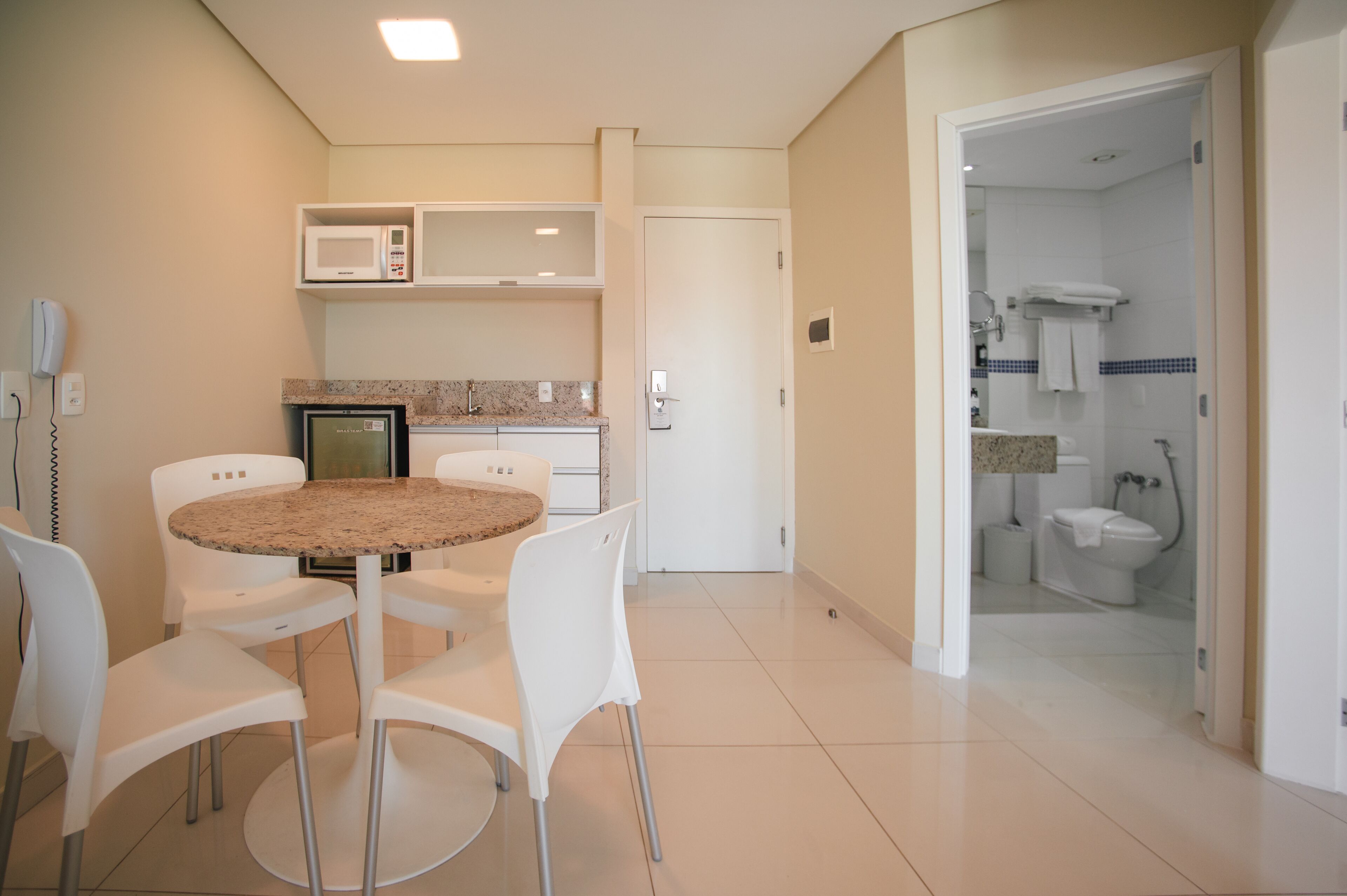 A hotel room with a dining area featuring a round table and four chairs, a microwave, a refrigerator, and a door leading to a bathroom with a toilet and sink.