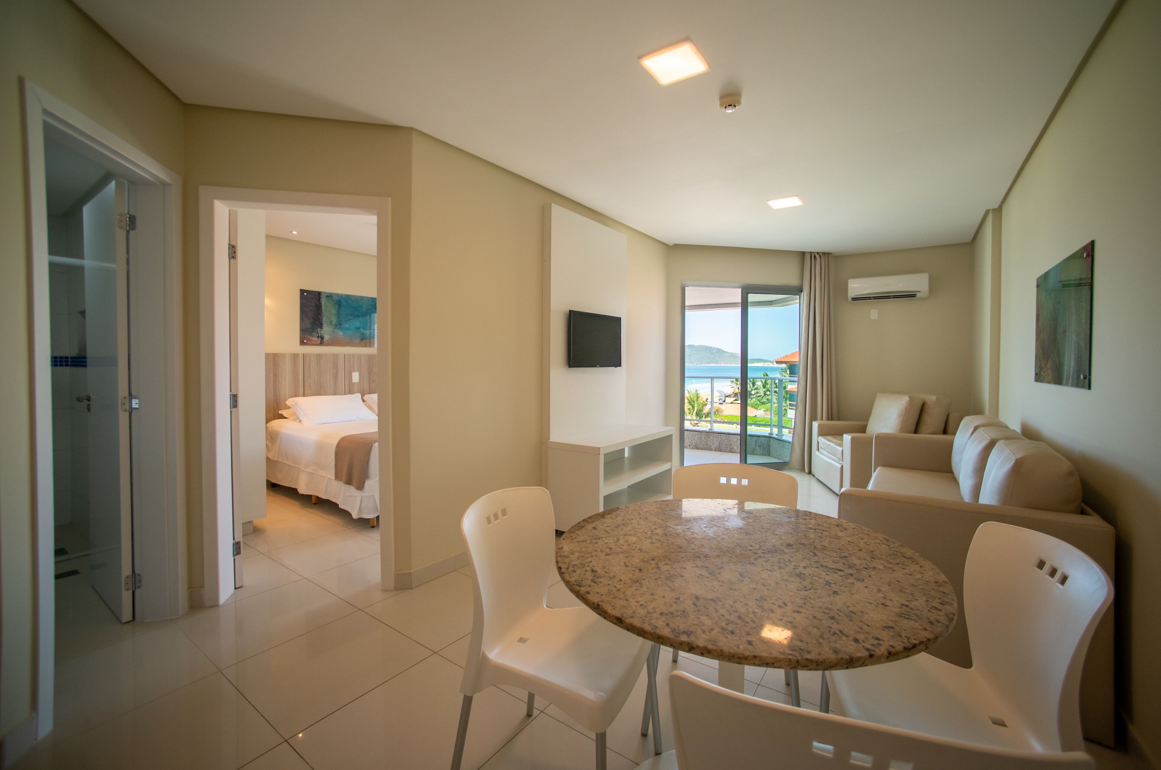 A hotel room with a bed, a dining table with chairs, a sofa, a TV, and a balcony with a view.