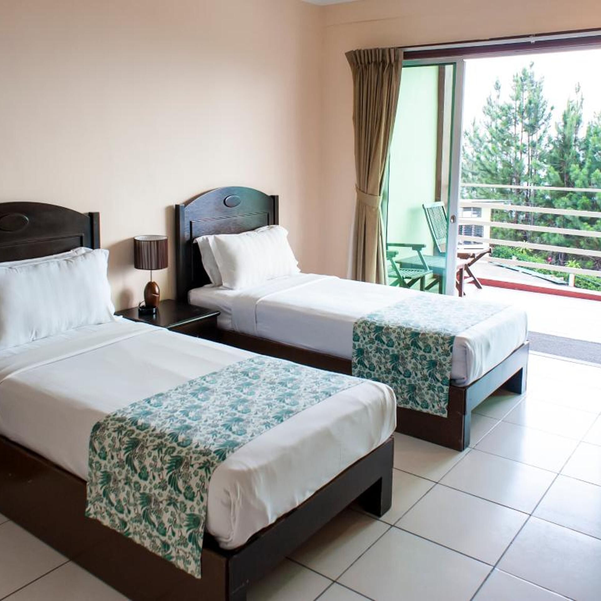 A hotel room with two beds, a nightstand, a lamp, and a view of the outdoors.