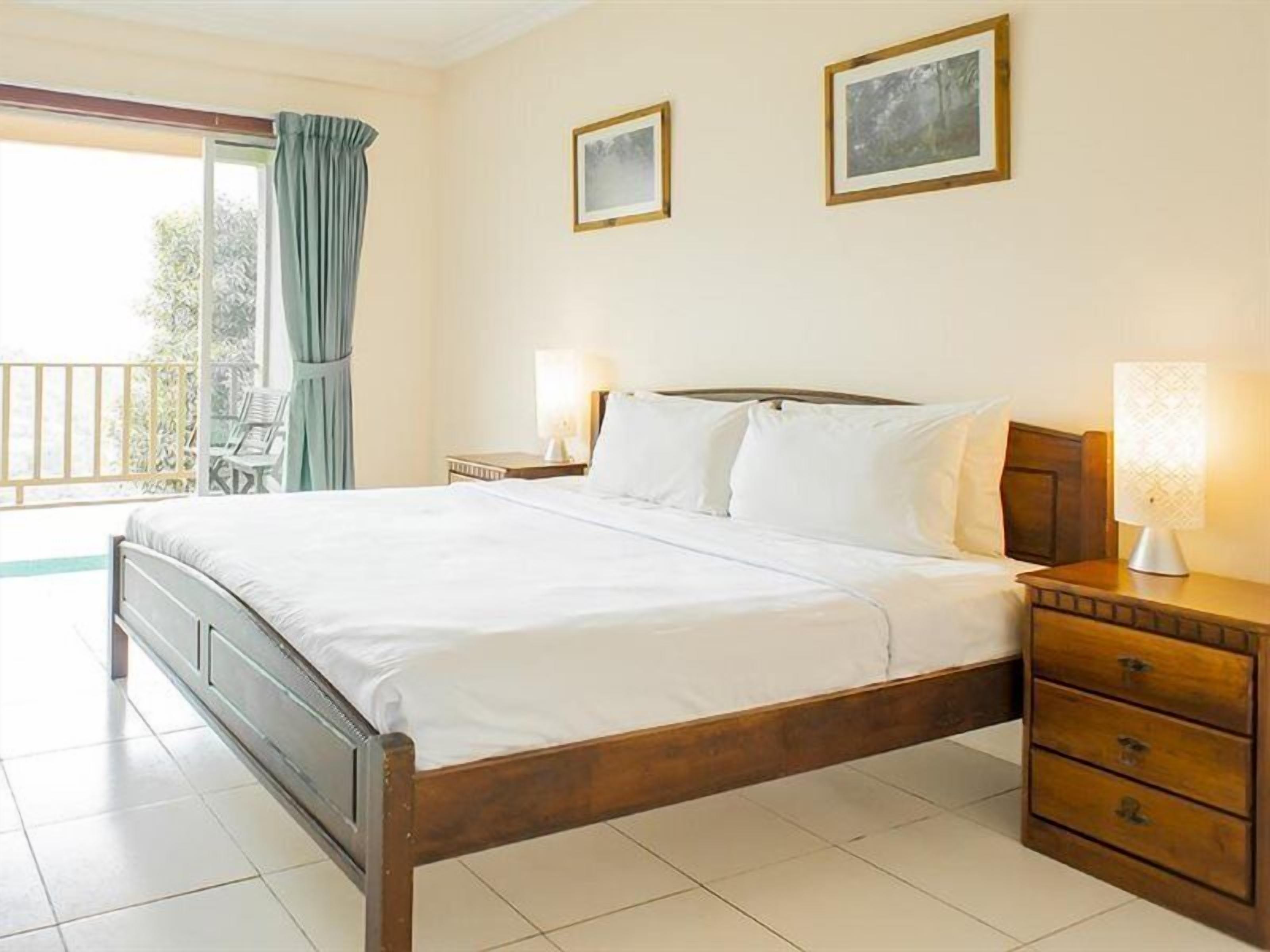 A neatly made bed in a room with a balcony, a nightstand with a lamp, and two framed pictures on the wall.