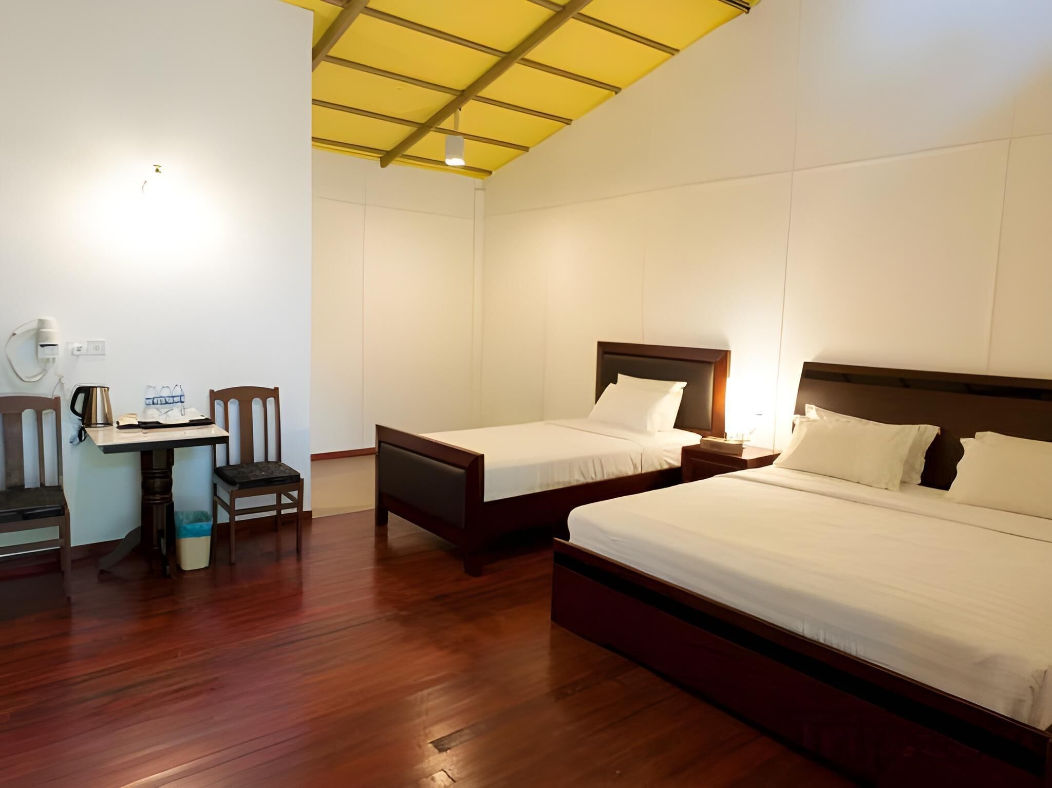 A hotel room with two single beds, a desk with a chair, a small table with a kettle, and a trash bin.