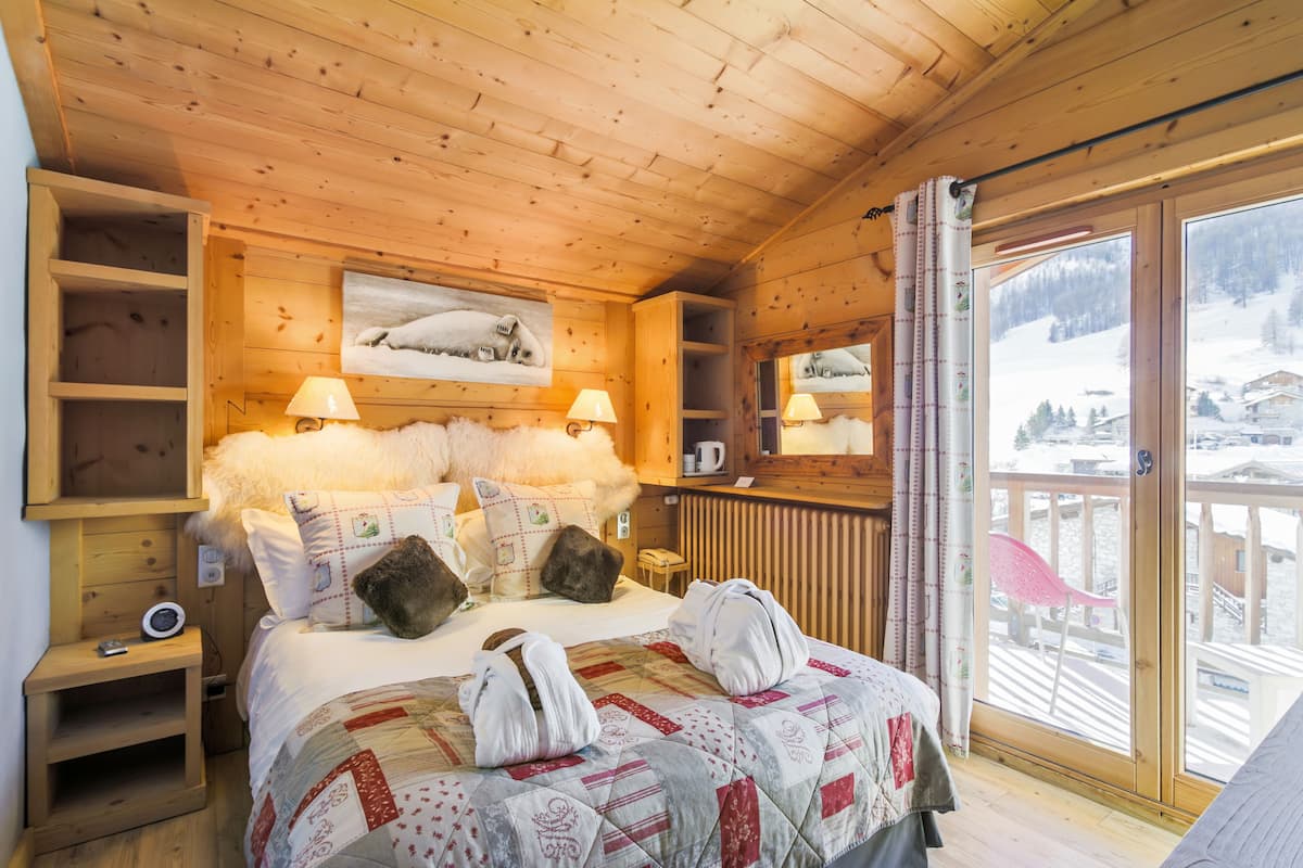 A cozy wooden cabin room with a bed, pillows, a nightstand, a mirror, and a window with a view of a snowy landscape.