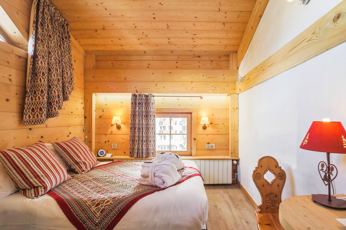 A wooden cabin room with a bed, pillows, a red lamp, and a patterned blanket.