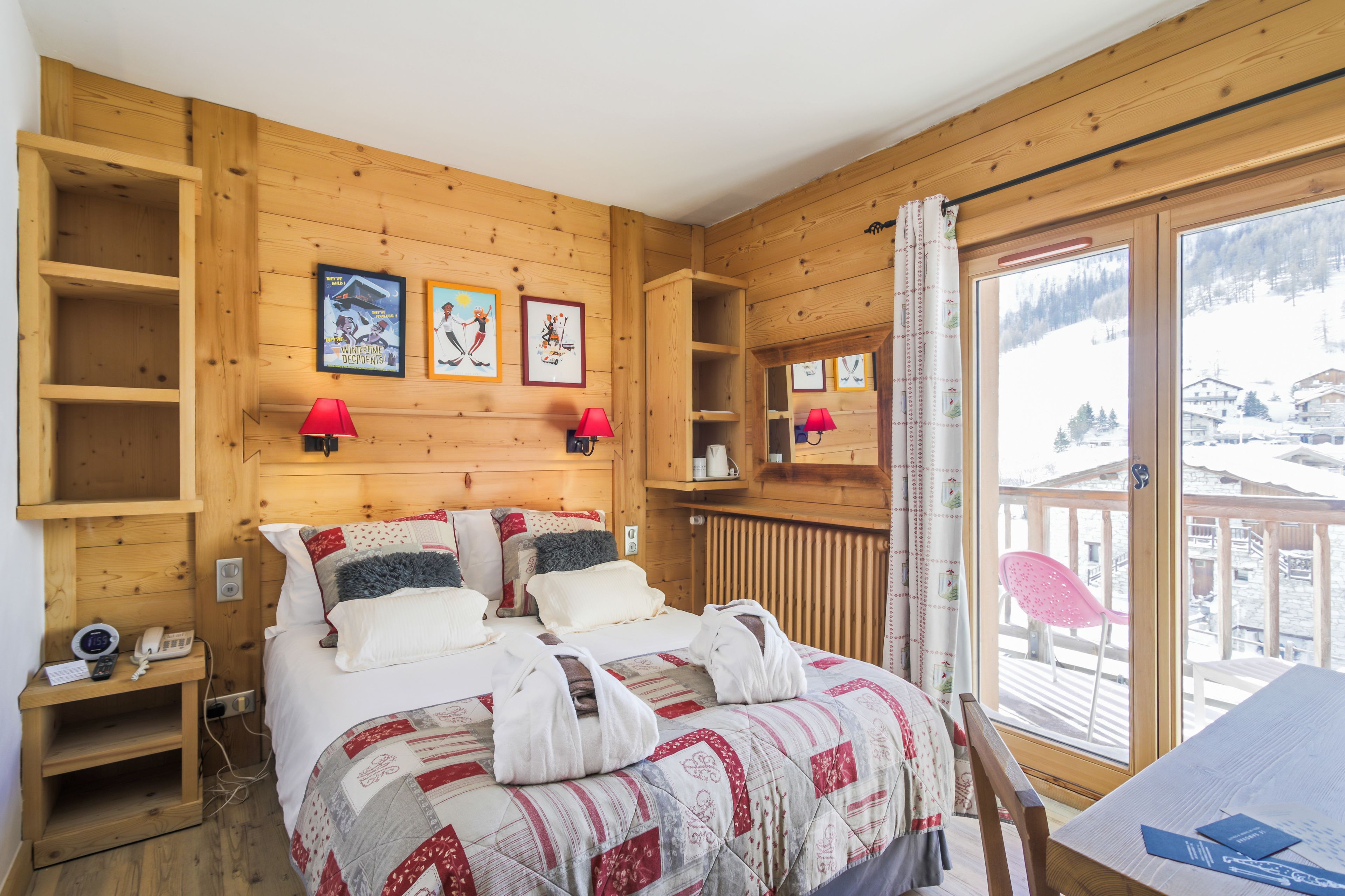 A wooden cabin room with a bed, two red lamps, a wooden shelf, and a window with a view of a snowy landscape.