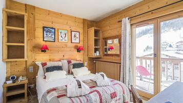 A wooden cabin room with a bed, two red lamps, a wooden shelf, and a window with a view of a snowy landscape.