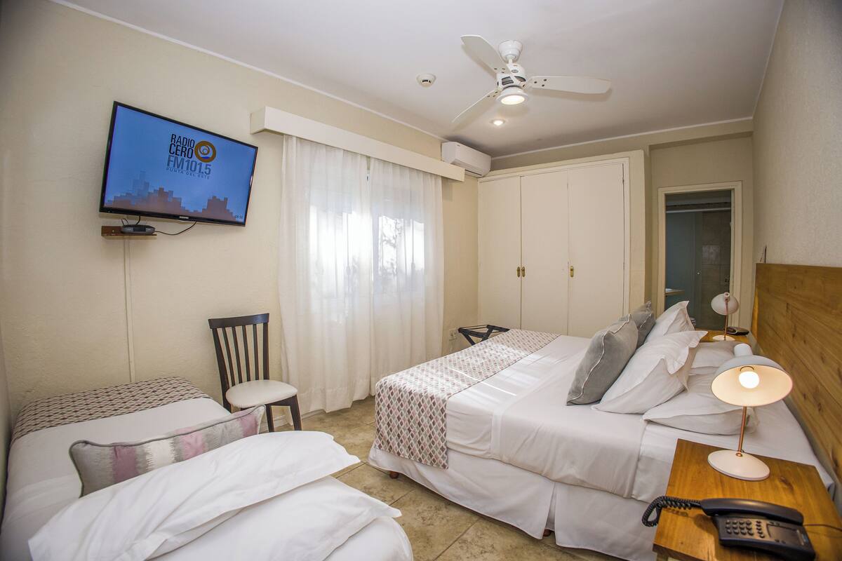 Habitación de hotel con cama, una silla, un escritorio con lámpara, teléfono, televisor montado en la pared y ventilador de techo.