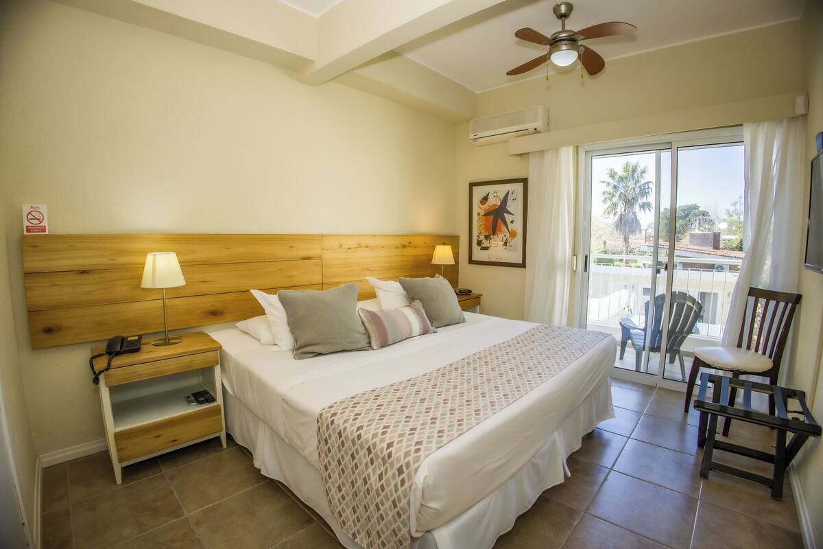 Habitación de hotel con cama, dos mesitas de noche, ventilador de techo, ventana con cortinas y una silla.