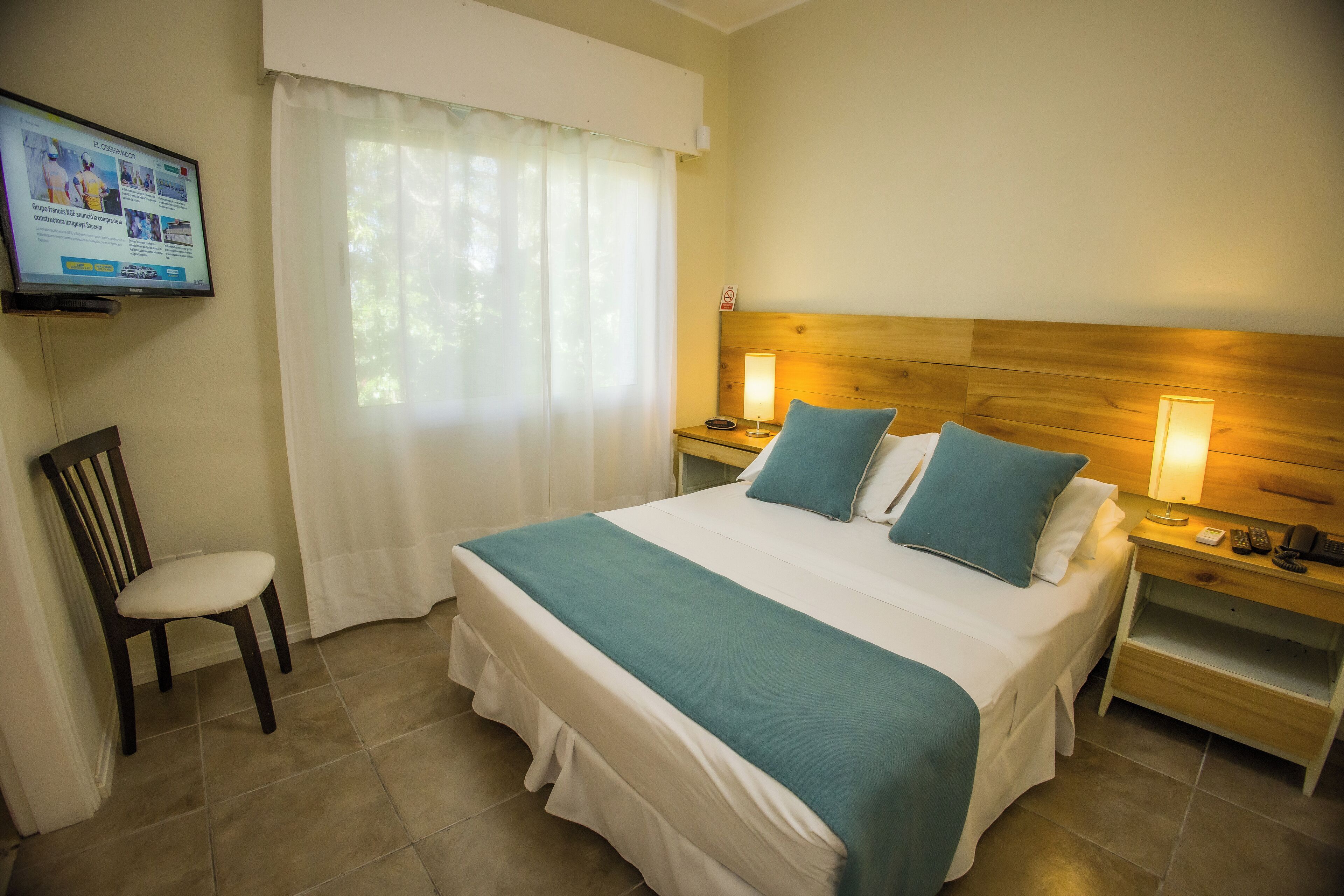 Habitación de hotel con cama, dos mesitas de noche, una silla, televisor montado en la pared y ventana con cortinas.