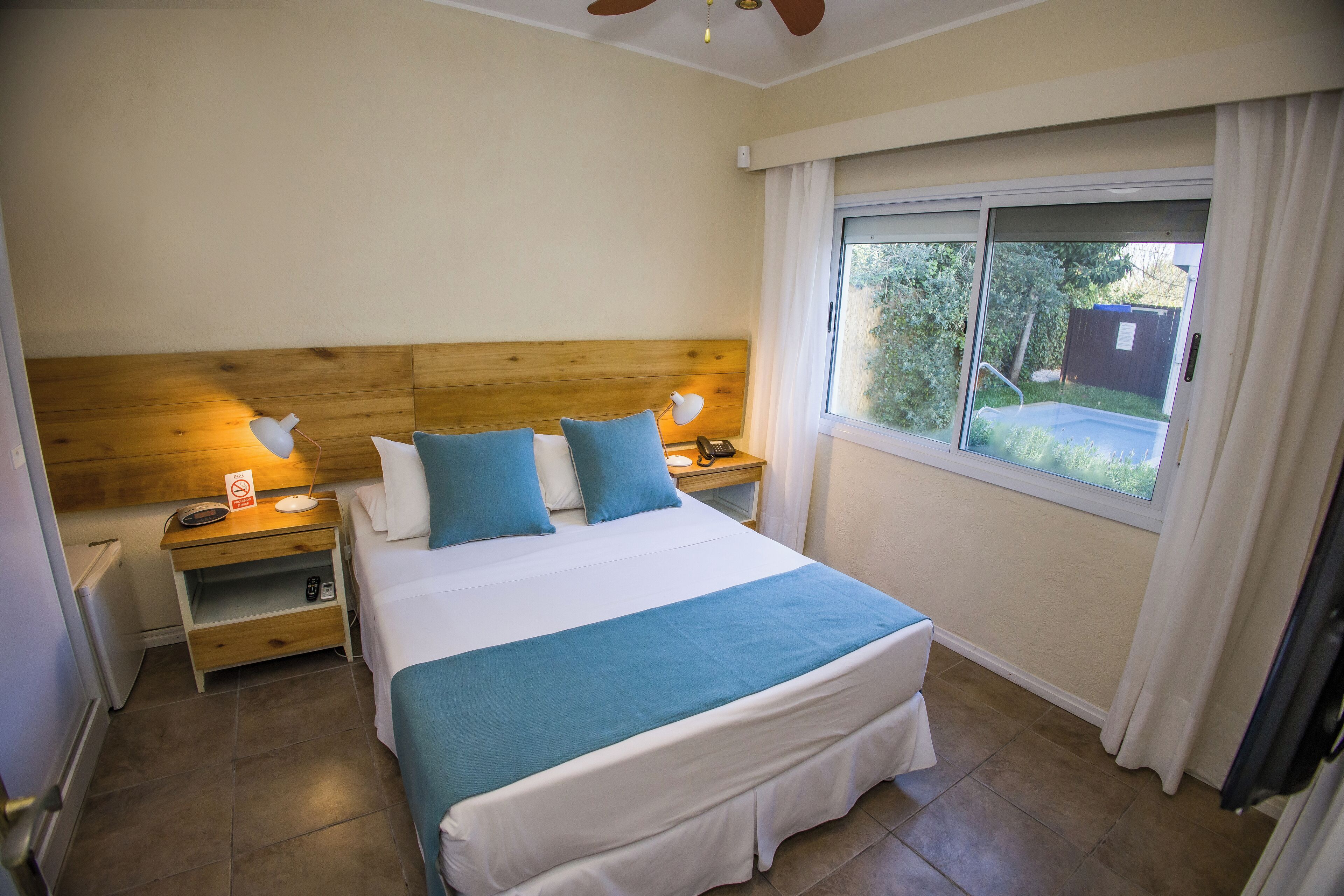 Un dormitorio con cabecera de madera, una cama grande con ropa de cama azul y blanca, mesitas de noche con lámparas, un calentador pequeño y una ventana con cortinas.