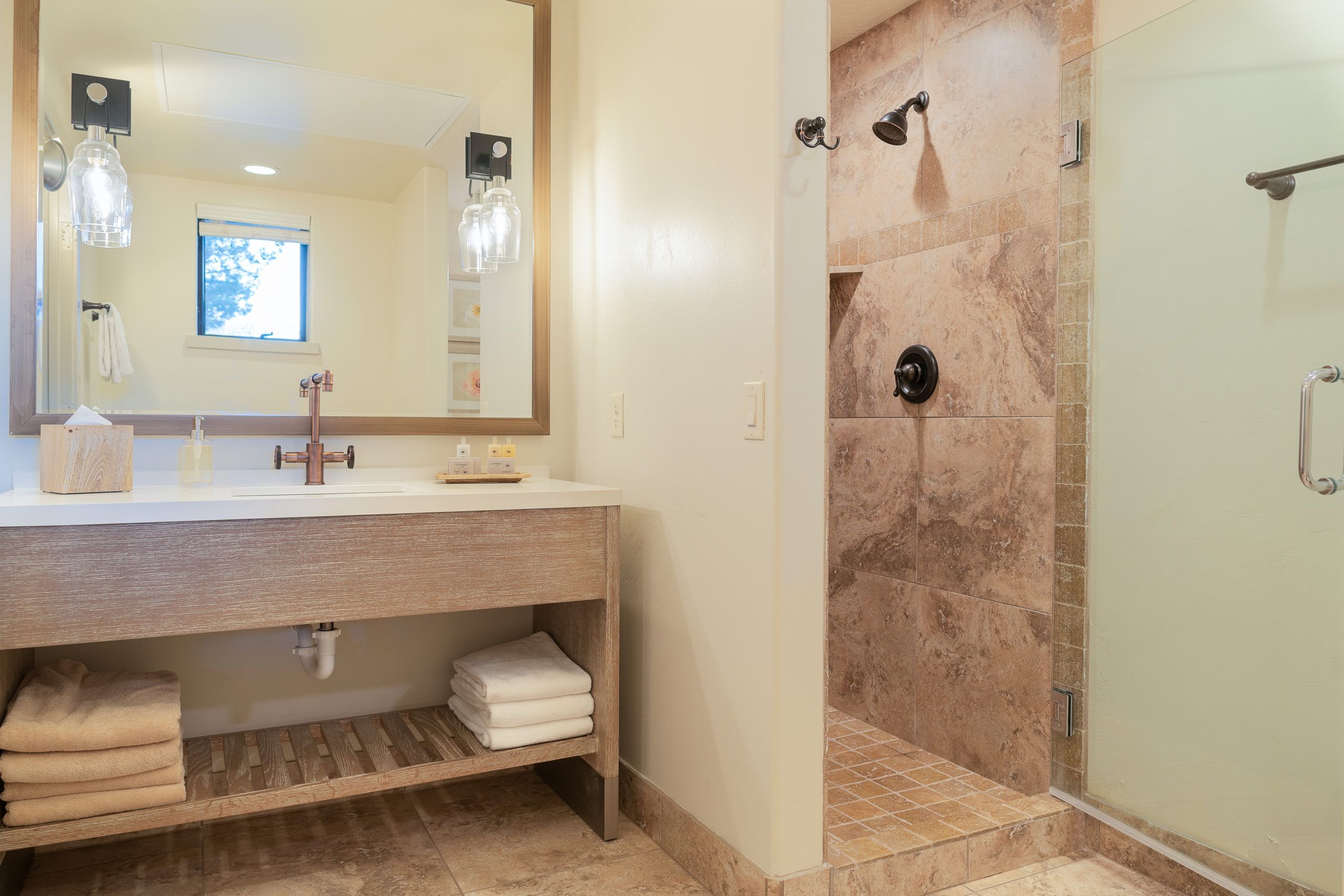 A bathroom with a wooden vanity, a large mirror, a shower with a glass door, and a window providing natural light.