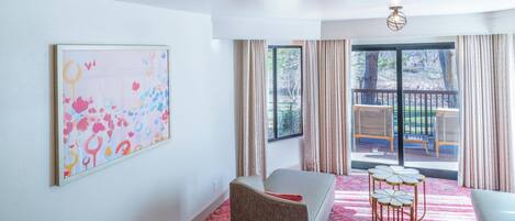 Habitación de hotel con un ventanal grande, un balcón con mobiliario exterior, un cuadro abstracto de colores y una alfombra roja.