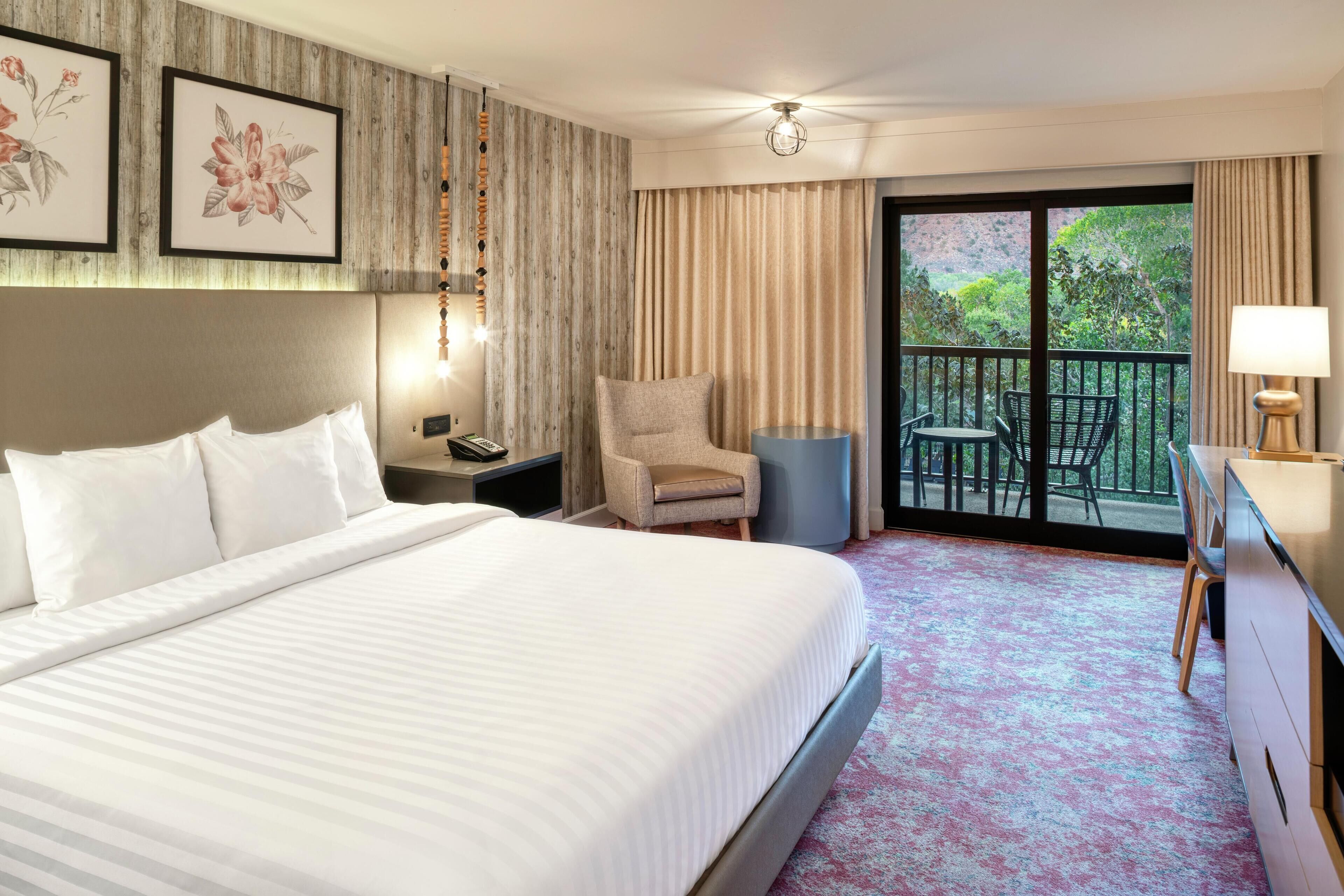 A hotel room with a large bed, a chair, a desk, and a balcony with a view of greenery.