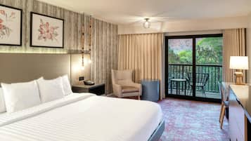 A hotel room with a large bed, a chair, a desk, and a balcony with a view of greenery.