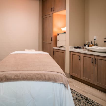 A room with a massage table, wooden cabinets, a sink, and a mirror.