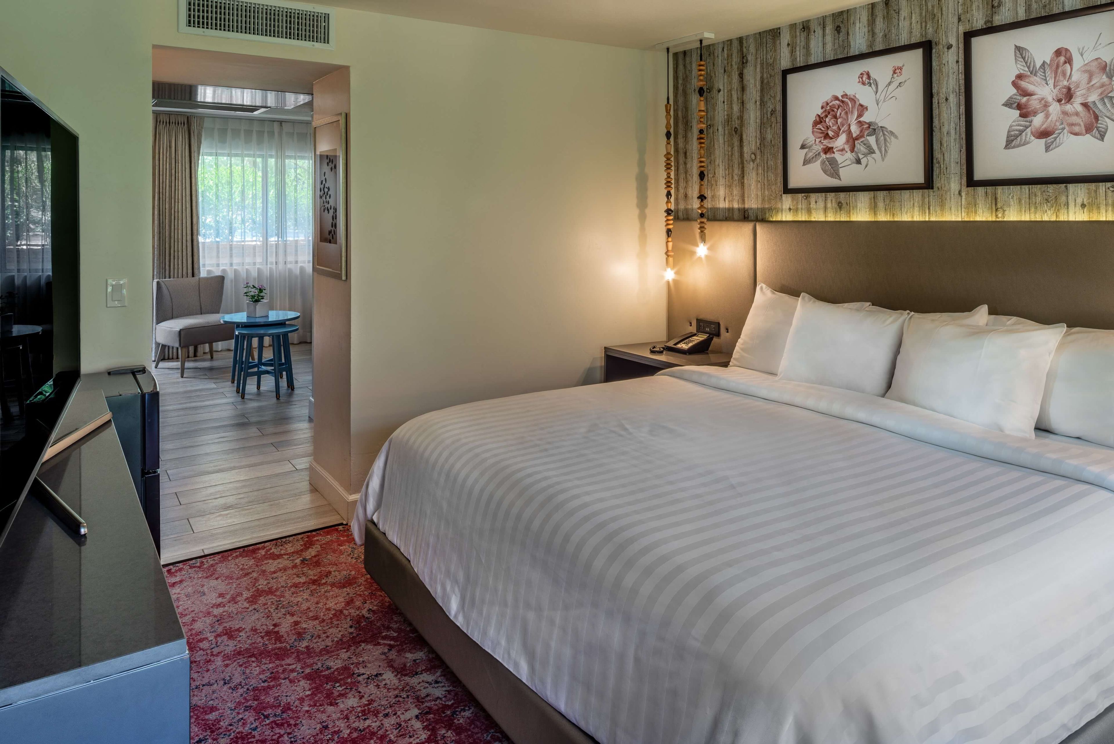A hotel room with a large bed, a desk with a computer, a chair, a small table with a plant, and a nightstand with a lamp.