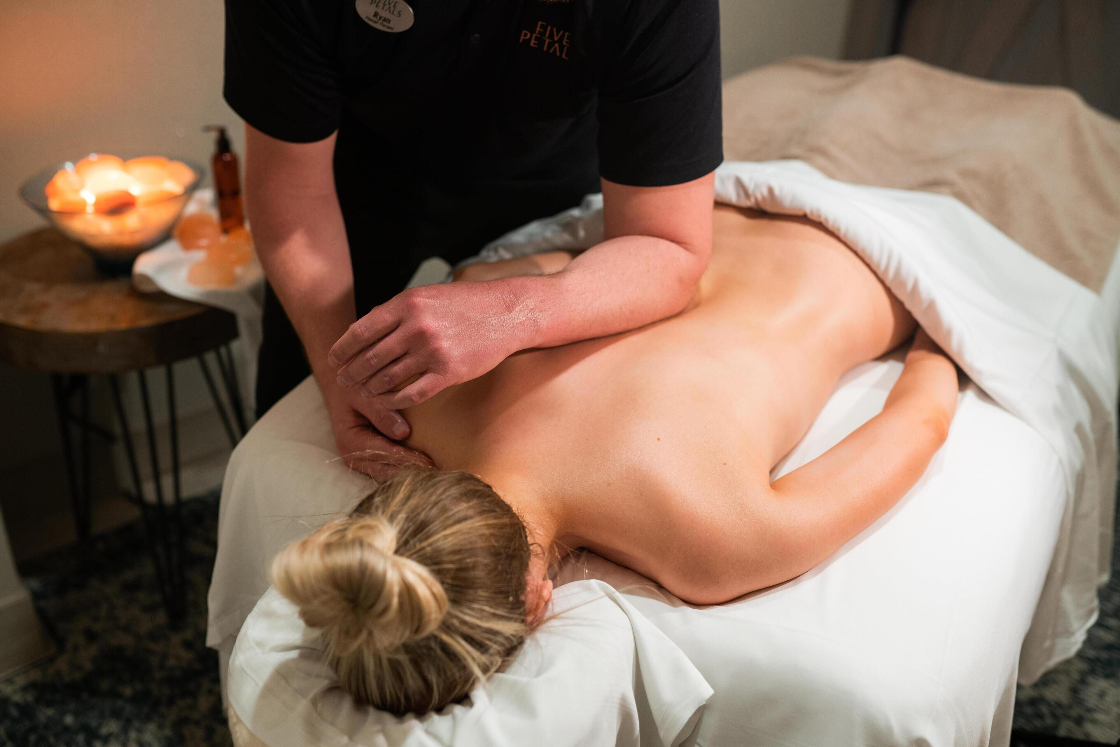 A massage therapy session in progress with a client lying face down on a massage table, a therapist's hands on the client's back, and a bowl of candles in the background.