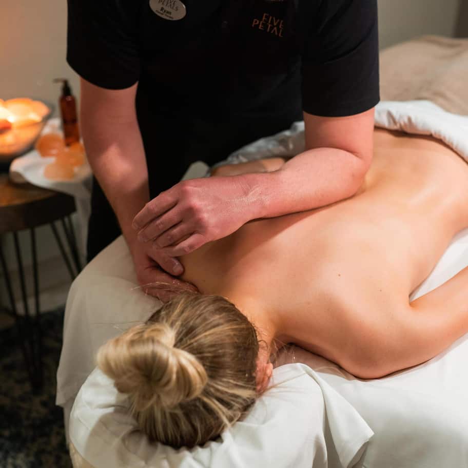 A massage therapy session in progress with a client lying face down on a massage table, a therapist's hands on the client's back, and a bowl of candles in the background.