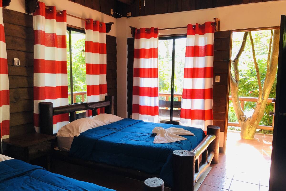 A room with two beds, red and white striped curtains, wooden headboards, and a view of greenery outside.