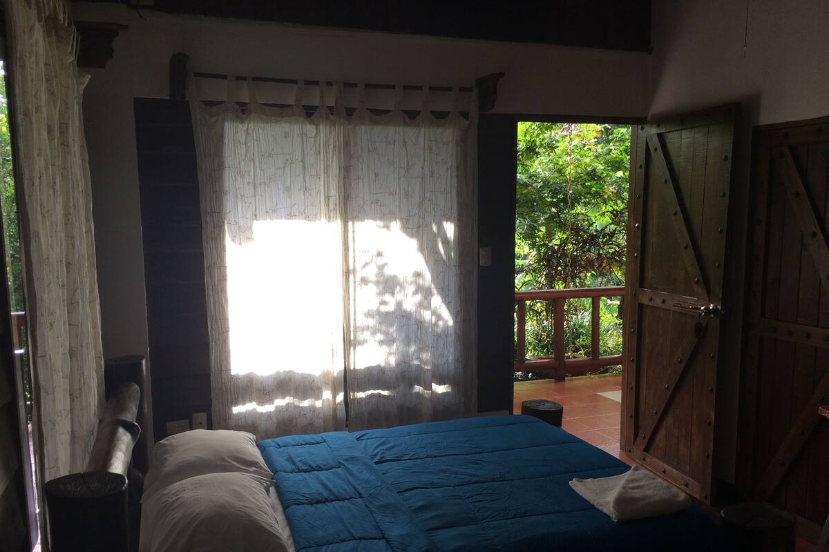 A room with a bed, a window with sheer curtains, and a view of greenery outside.