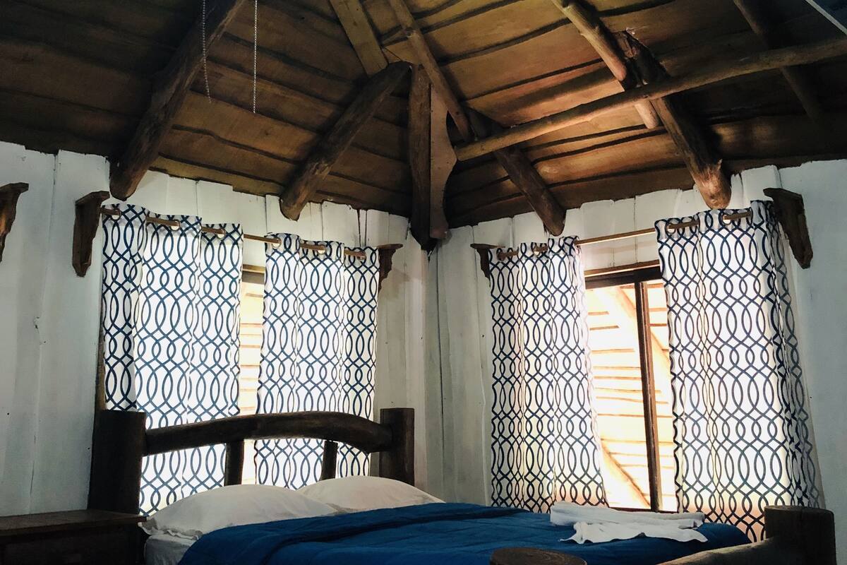 A room with a wooden ceiling, two beds with blue bedding, and patterned curtains.