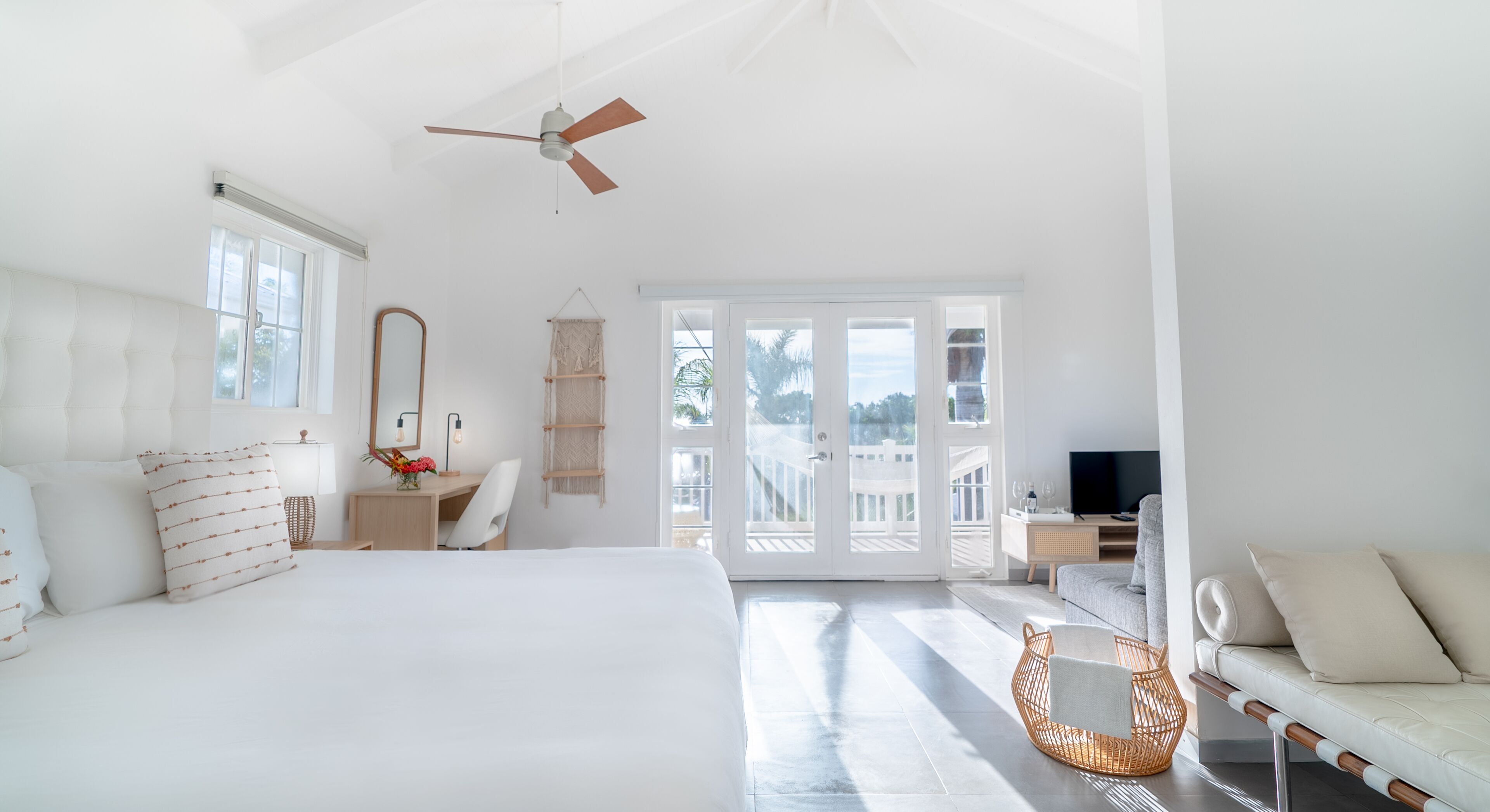 A spacious bedroom with a large bed, a desk, a chair, a wicker chair, a sofa, a TV, and a balcony with a view.