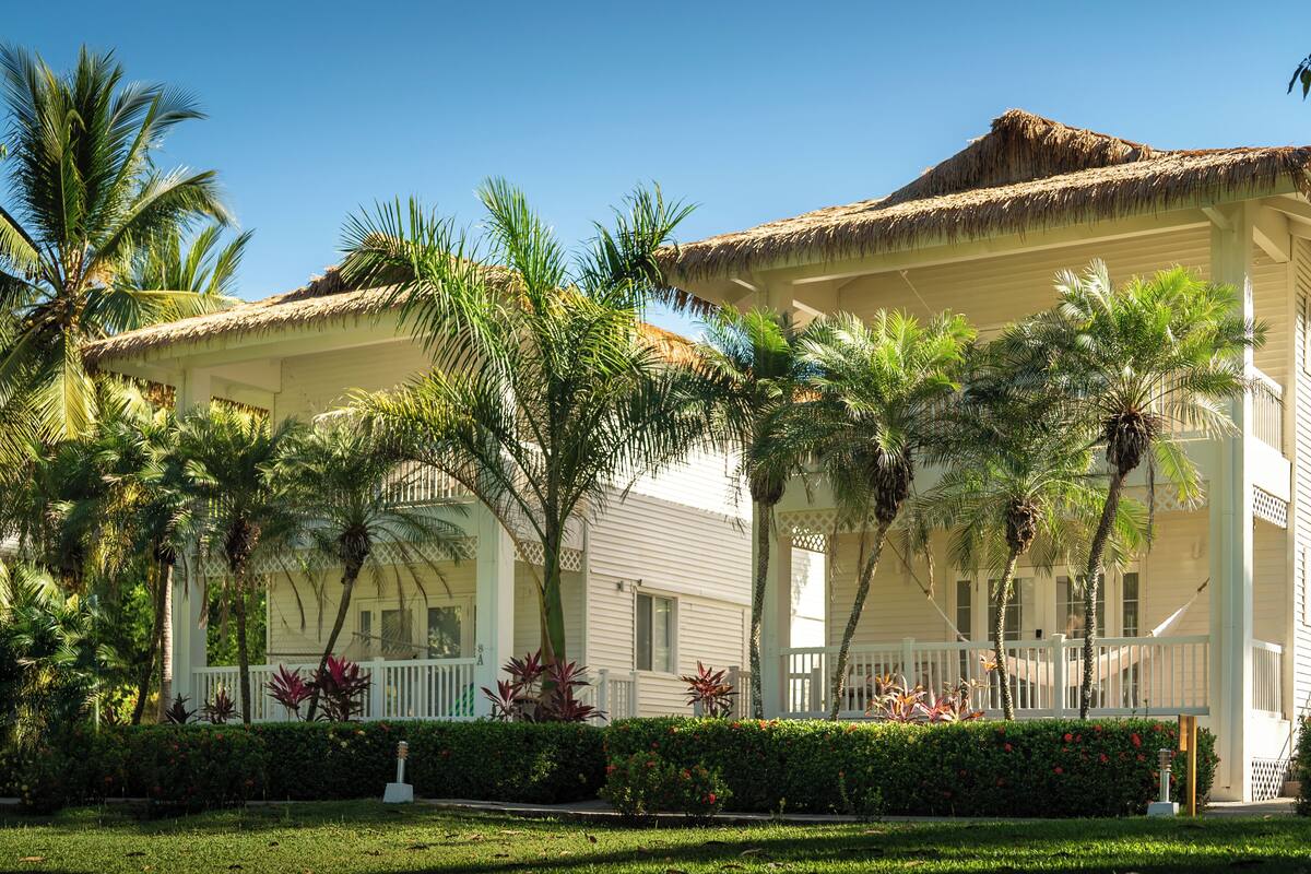 A white building with a thatched roof, surrounded by palm trees and a well-maintained garden.