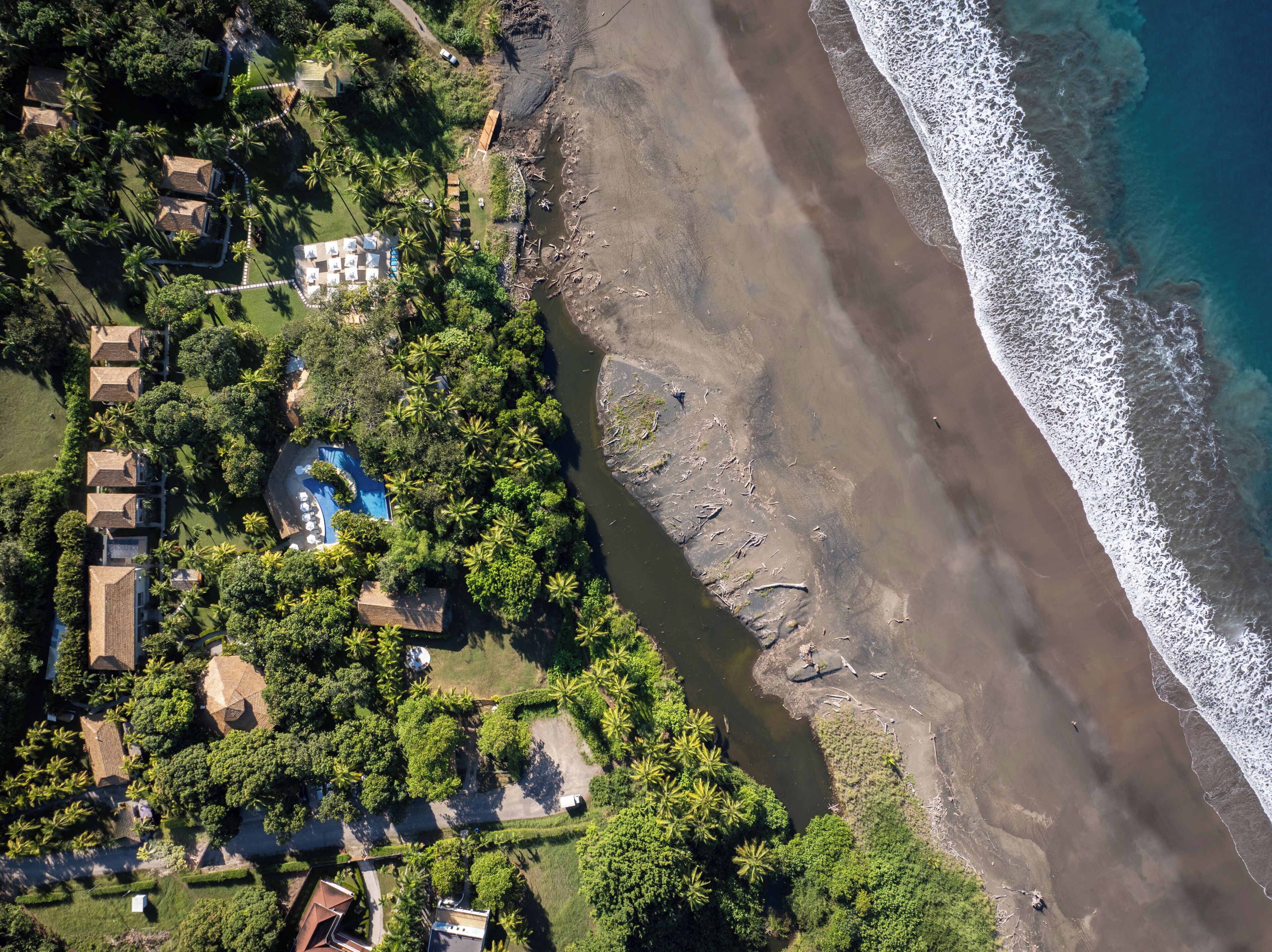 Ein Luftbild einer Küstenregion mit Strand, einem von üppigem Grün umgebenen Schwimmbad und mehreren Gebäuden mit unterschiedlichen Dachtypen.