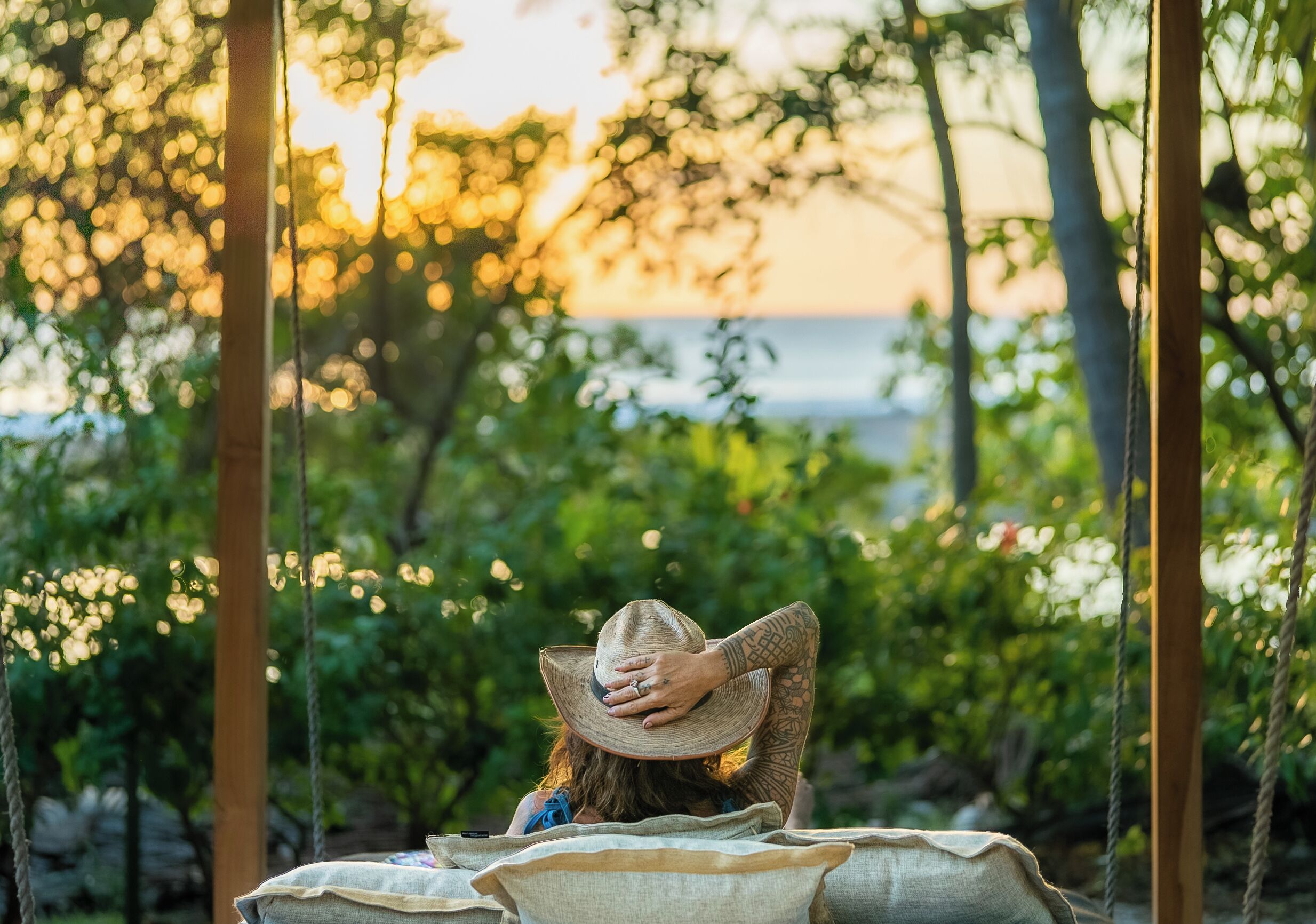 A person sitting on a swing with a hat, surrounded by greenery and a sunset in the background.