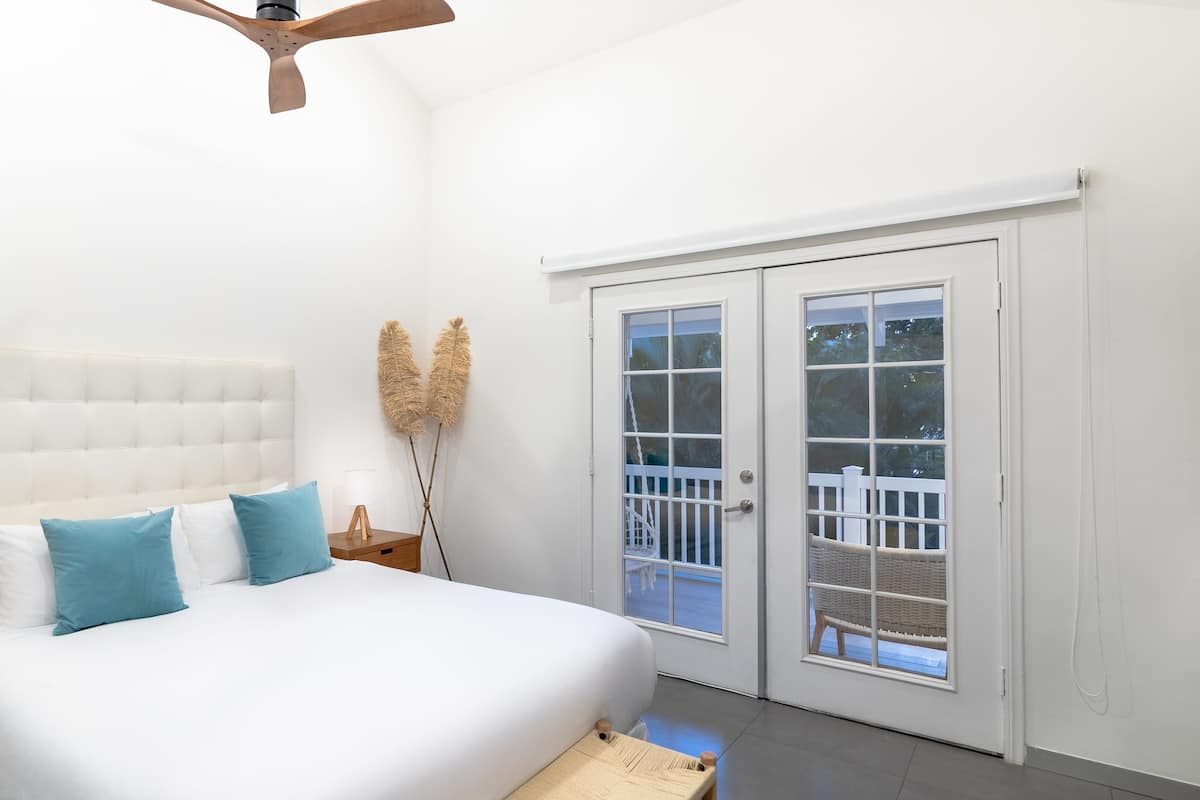 A bedroom with a tufted headboard, a bed with white and blue bedding, a wooden nightstand, a bedside lamp, a ceiling fan, and a sliding glass door leading to a balcony with a white fence.