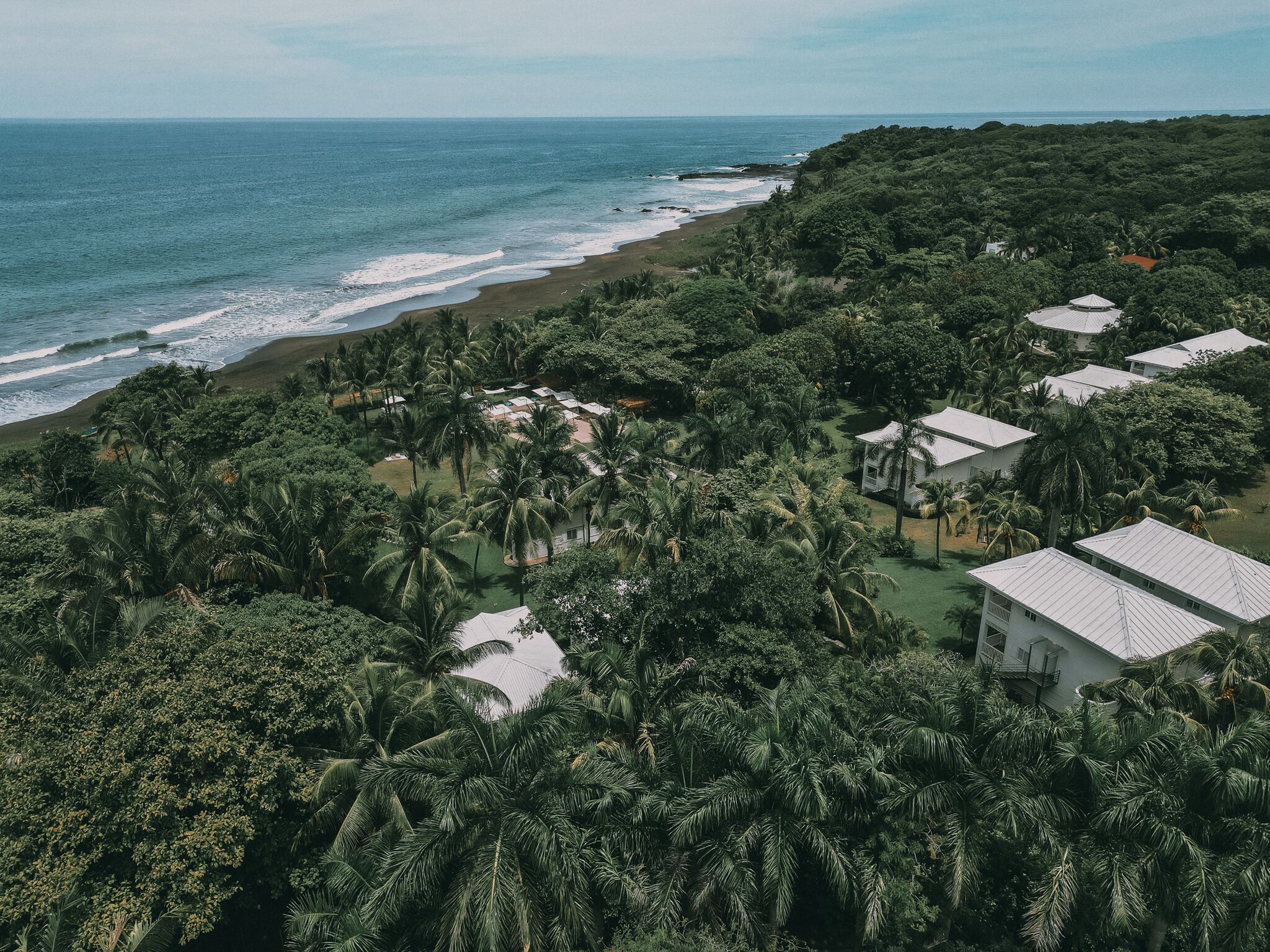 An aerial view of a coastal resort with multiple bungalow-style buildings surrounded by dense tropical vegetation and a beach.