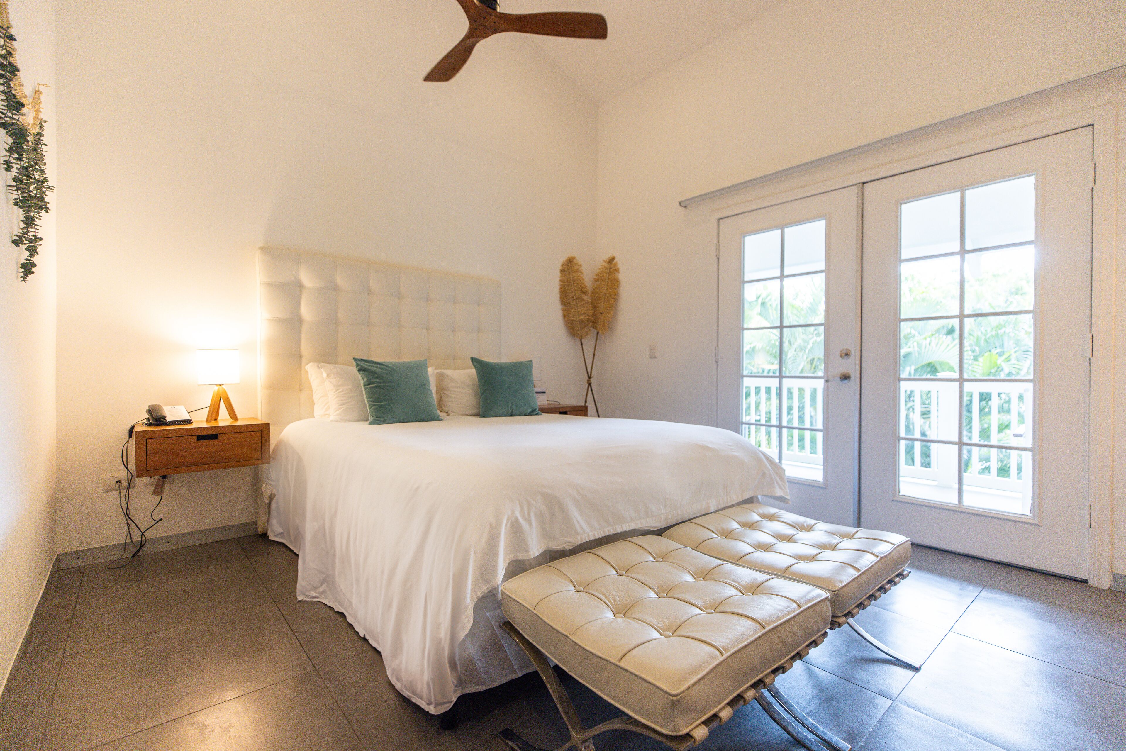 A bedroom with a bed, bedside table, lamp, and a view of greenery through the window.